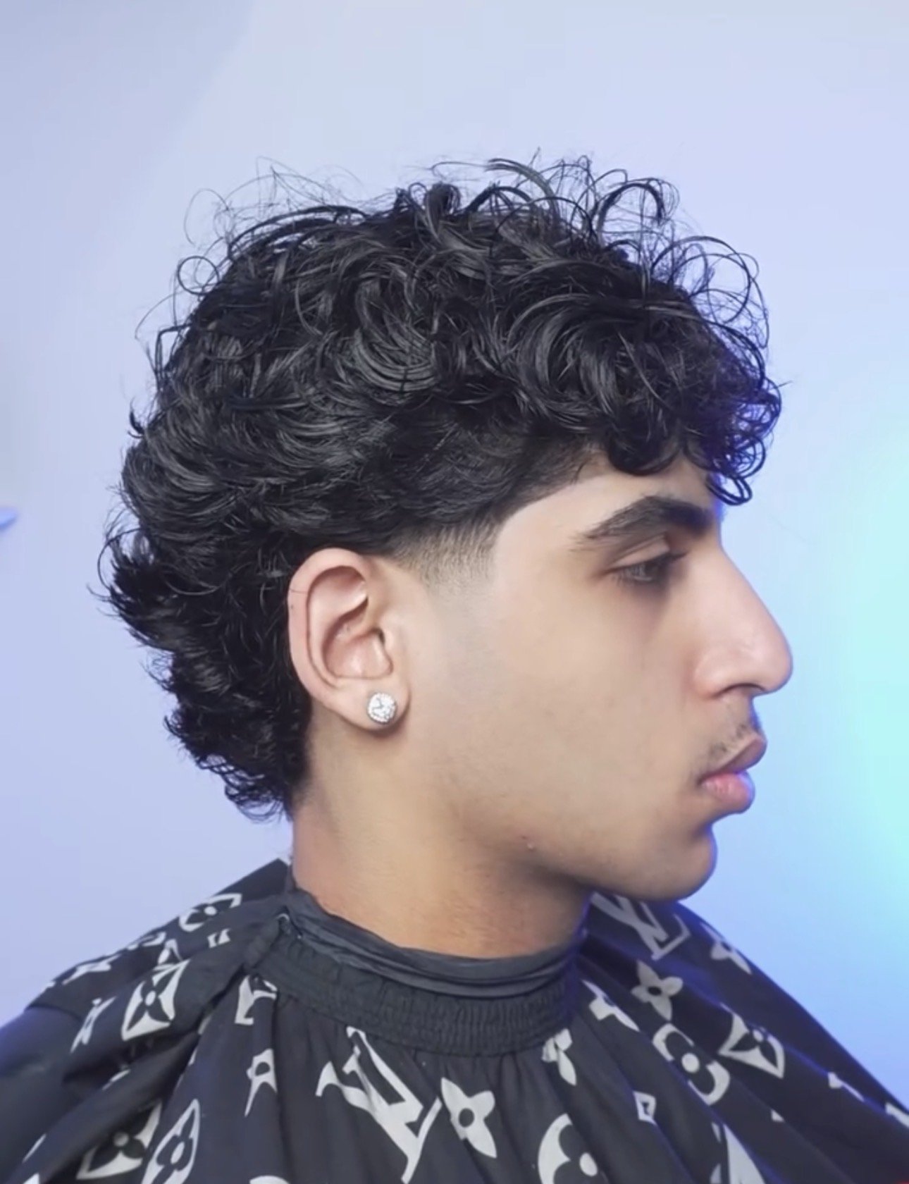 Side profile of a young man with black curly hair, wearing a diamond earring and a patterned barber cape, in a salon.