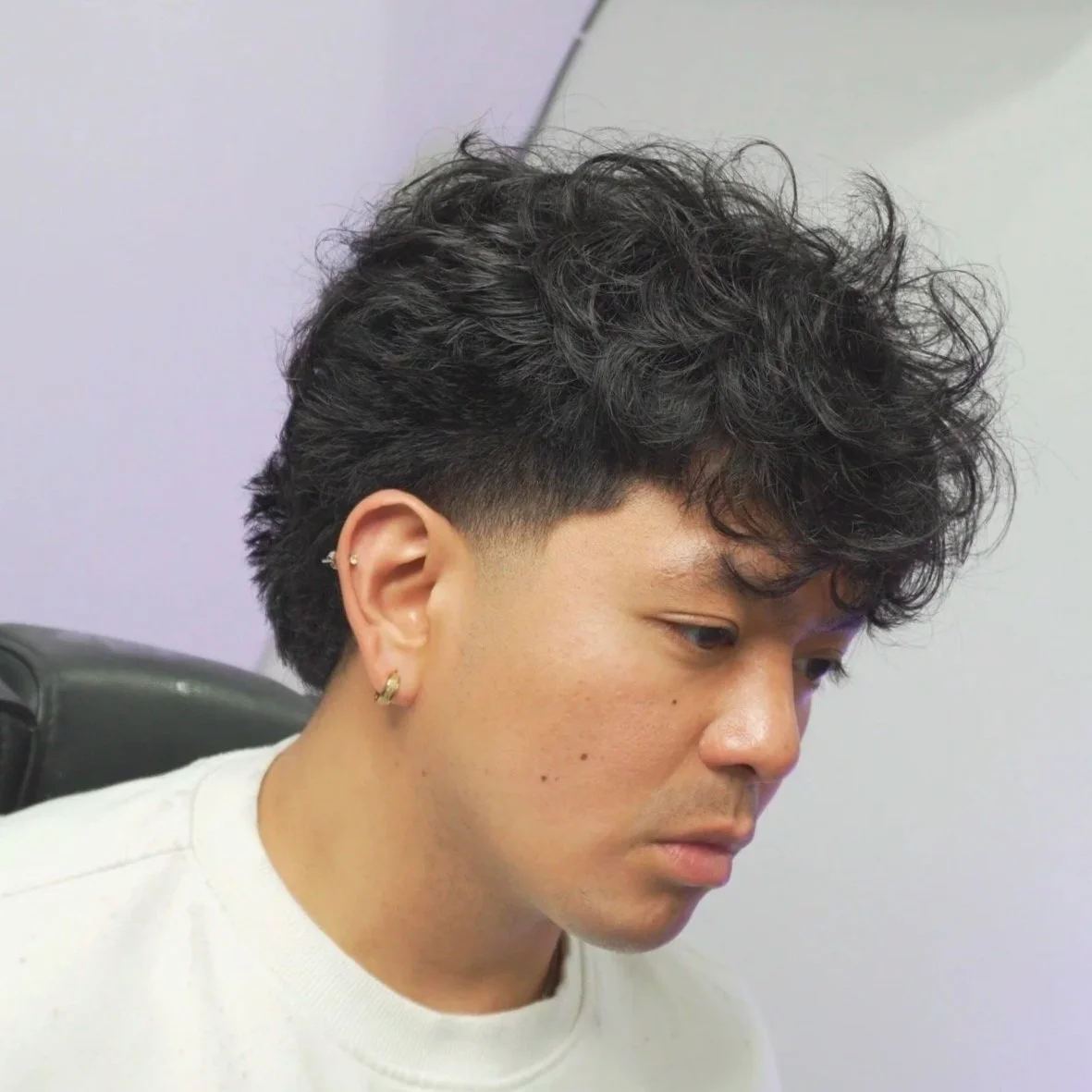 Close-up of a person with dark, curly hair styled in a high fade haircut, wearing earrings, looking down.