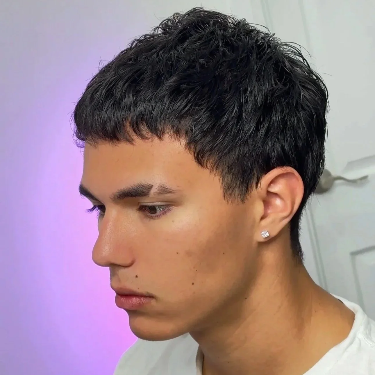 Profile of a young person with short, dark, textured hair, wearing a small earring, against a background with soft purple and white lighting.