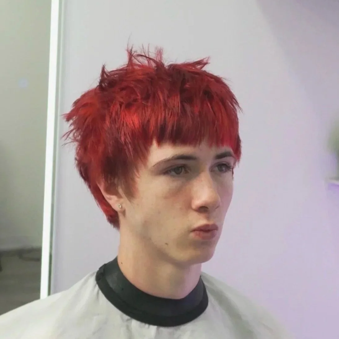 Young man with short, spiky, bright red hair sitting in a salon chair with a cape around her shoulders.