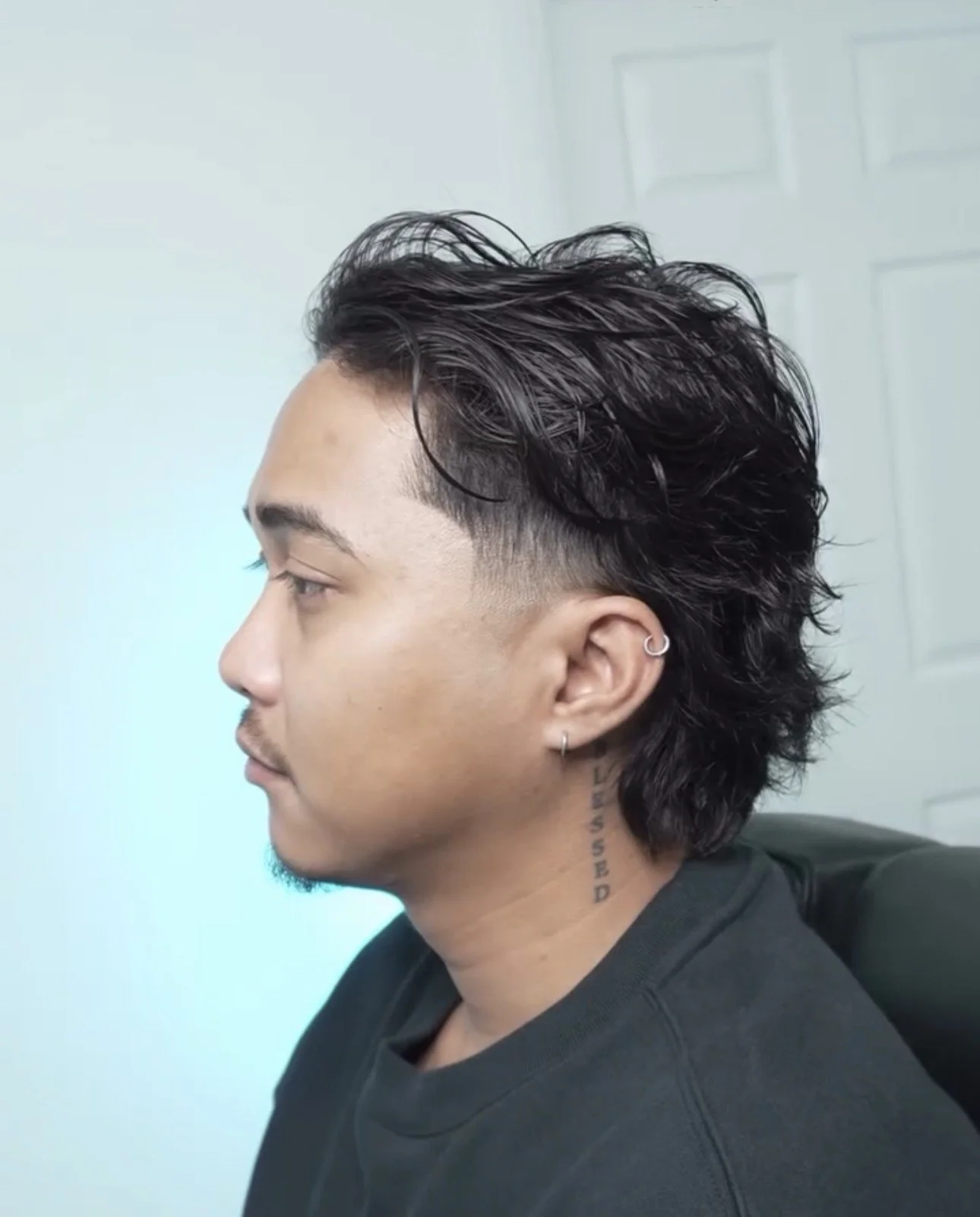 Side profile of a young man with dark, curly, and voluminous hairstyle, featuring a fade on the sides, wearing earrings, and a tattooed neck, sitting in a modern room.