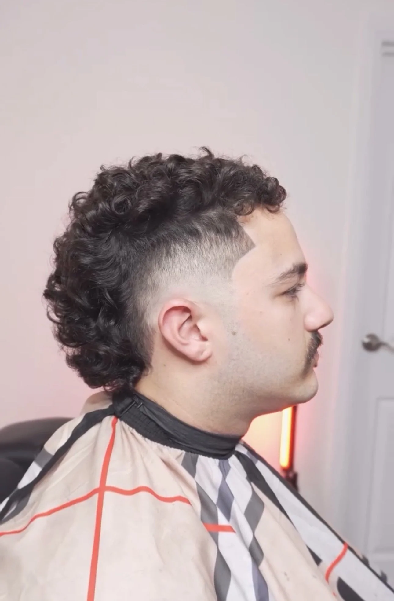 Man with curly dark hair and a shaved undercut sitting in a barber's chair, wearing a barber cape with red, black, and white geometric patterns, in a barber shop.