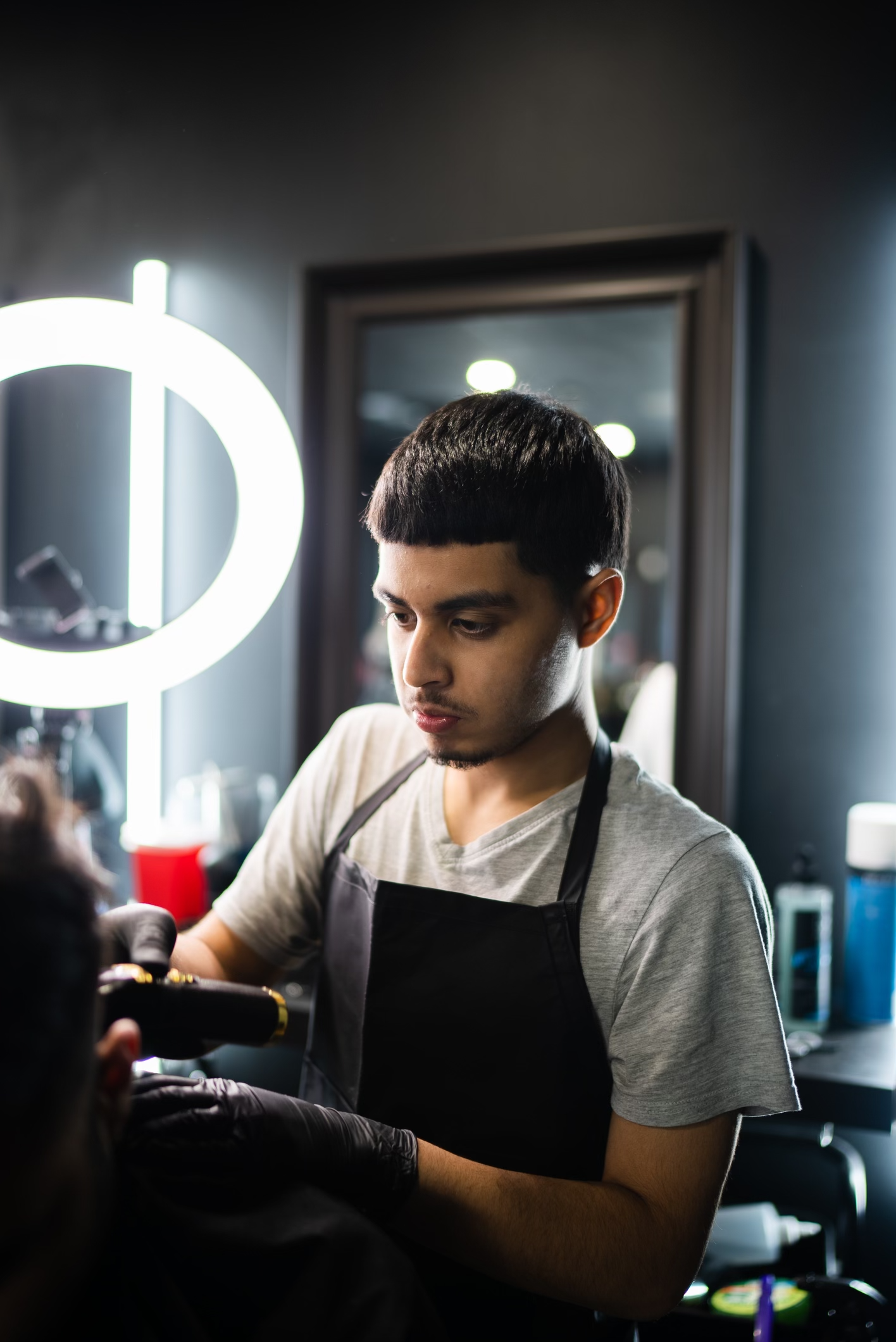 Barber with a short haircut, wearing a gray t-shirt and black apron, cutting a client's hair in a modern barber shop.