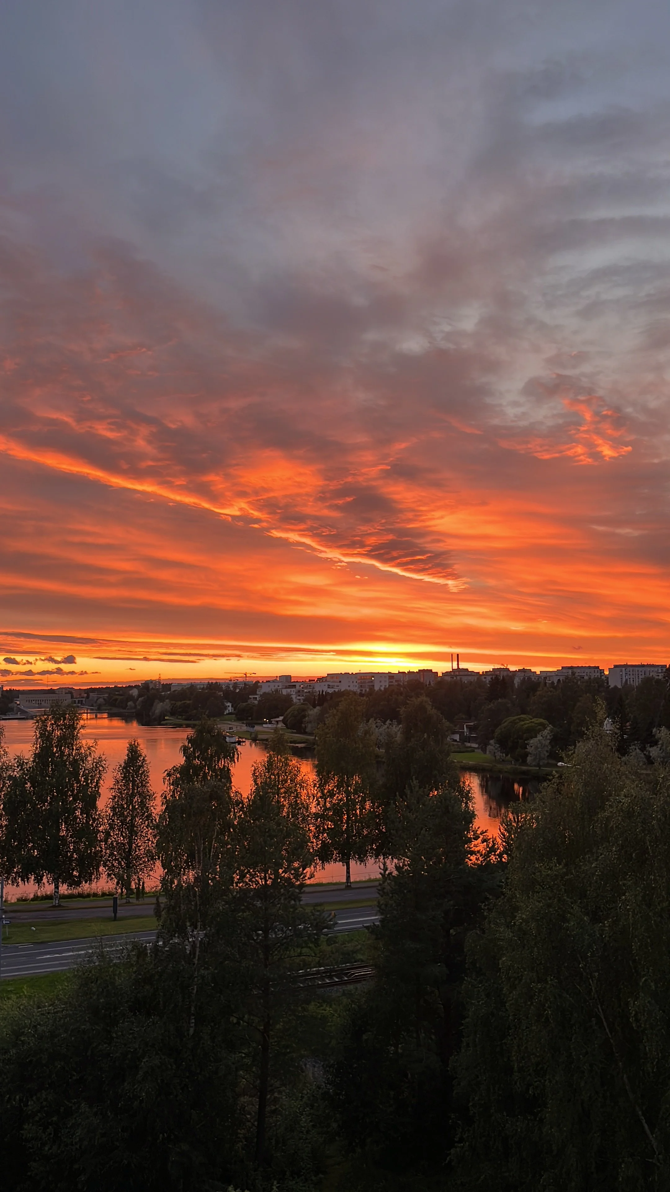 Sunset over a river with a partly cloudy sky and cityscape in the background. Trees are in the foreground.