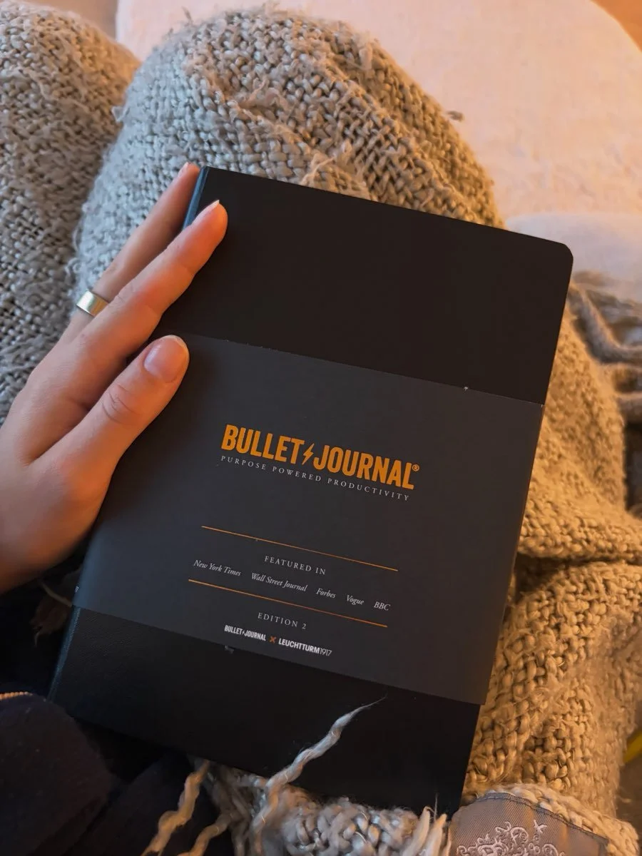 A person holding a black Bullet Journal notebook resting on their lap, with a textured beige and cream-colored blanket or pants nearby.