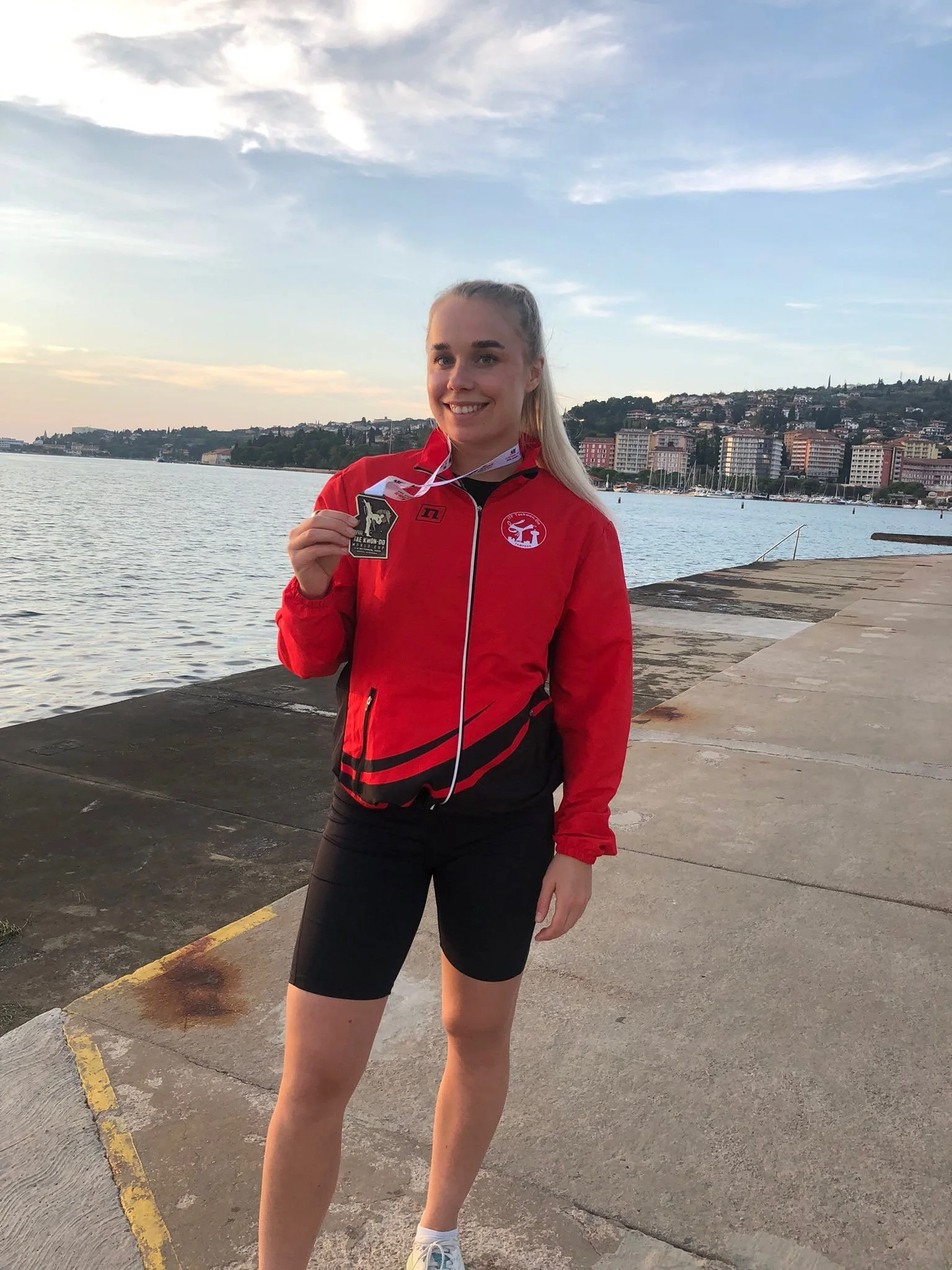 Young woman in athletic attire holding a medal by a waterfront with buildings and boats in the background.