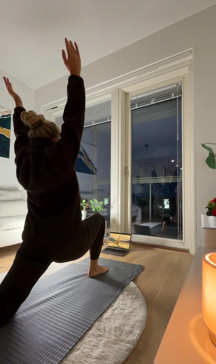 Woman practicing yoga in a lunge position on a yoga mat at home, with a laptop displaying a yoga instructor, in a living room at night.