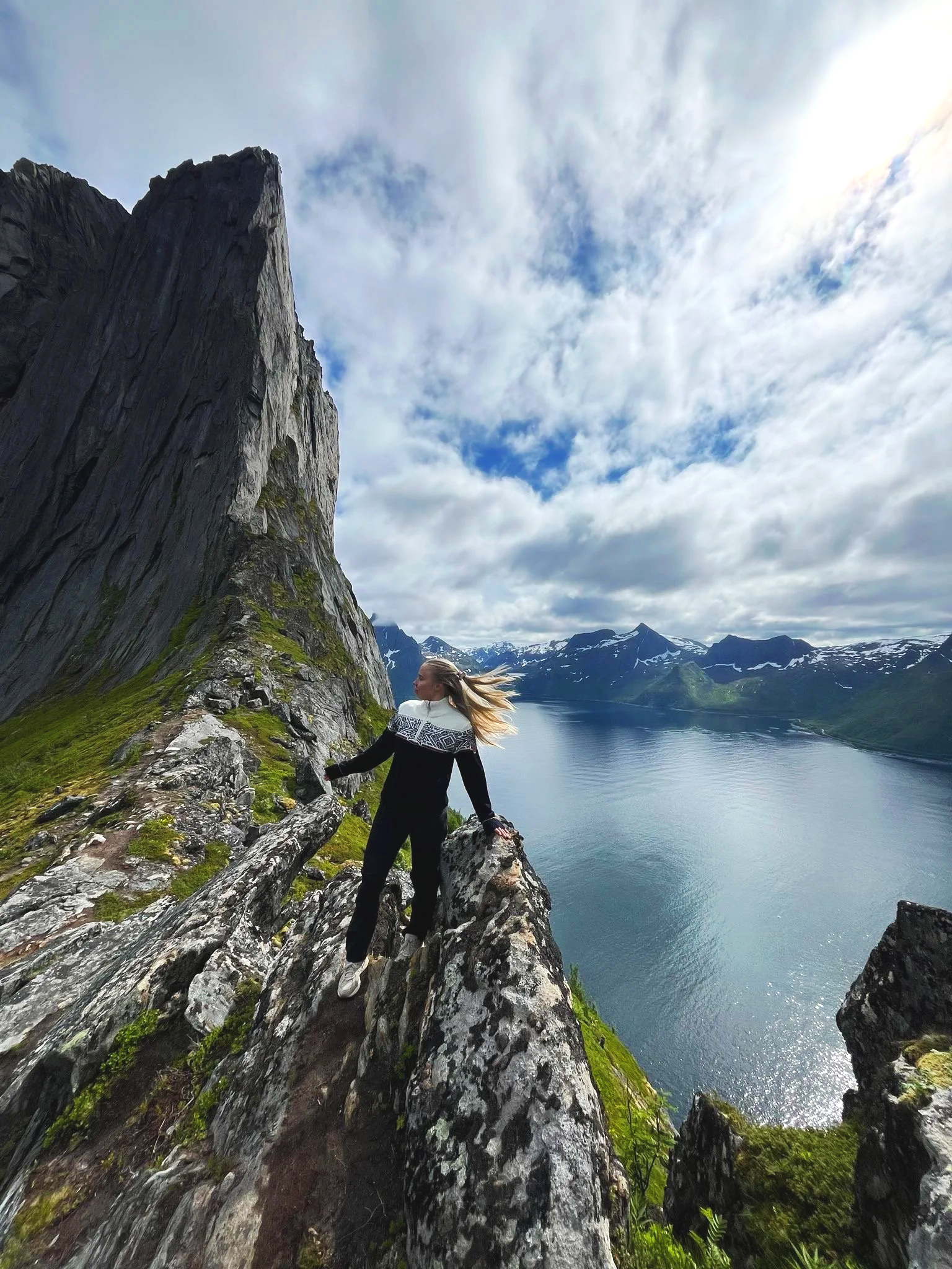 A woman dressed in black and white hiking gear standing on a rocky ledge on a mountain with a large cliff to her left, overlooking a fjord with snow-capped mountains in the distance under a partly cloudy sky.
