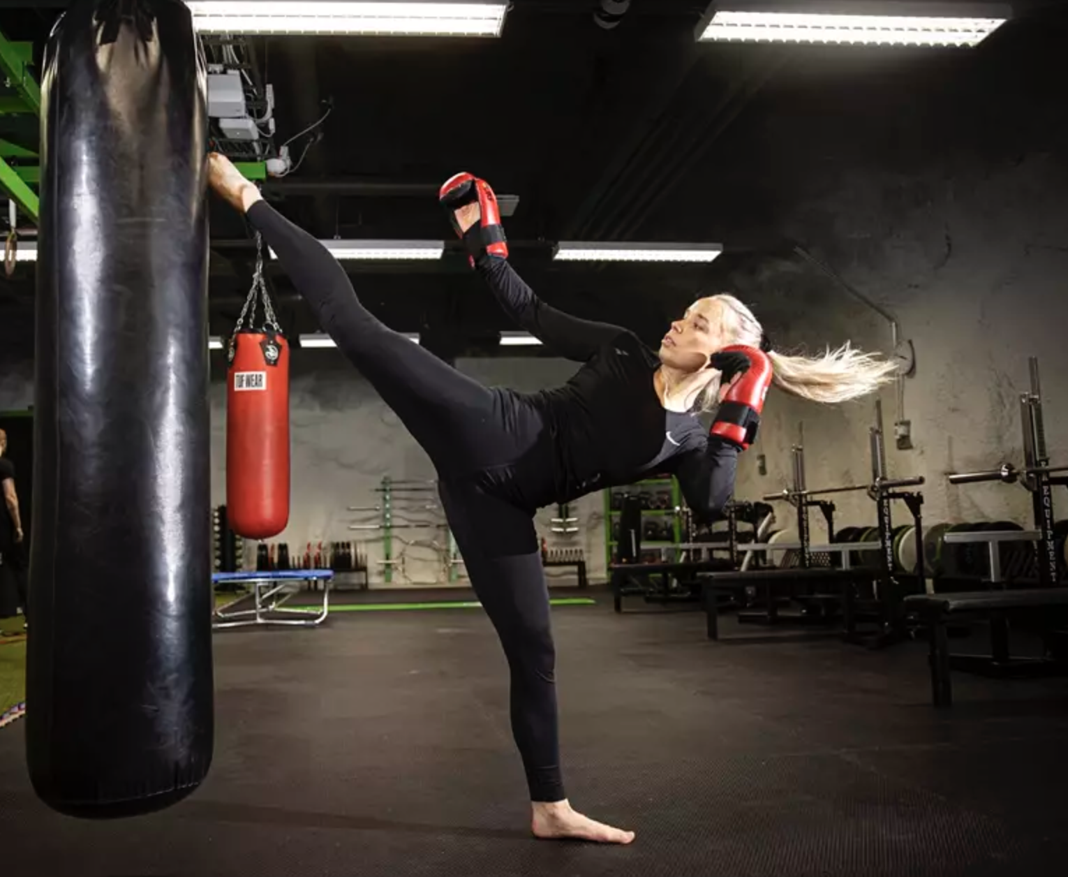 A woman practicing martial arts in a gym, executing a high kick towards a hanging punching bag while wearing red gloves and a black athletic outfit.