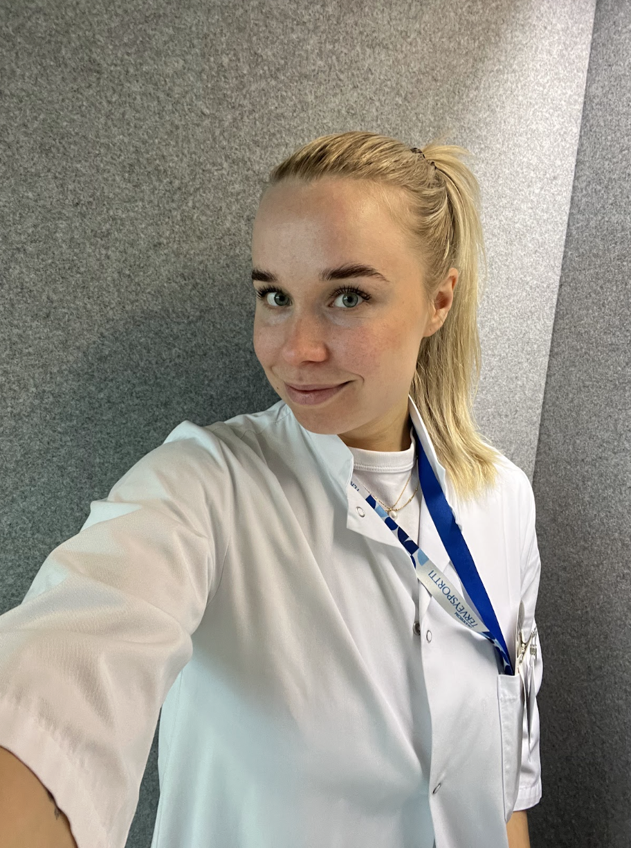 A young woman with blonde hair tied back, wearing a white medical coat and a blue lanyard, taking a selfie against a gray textured wall.