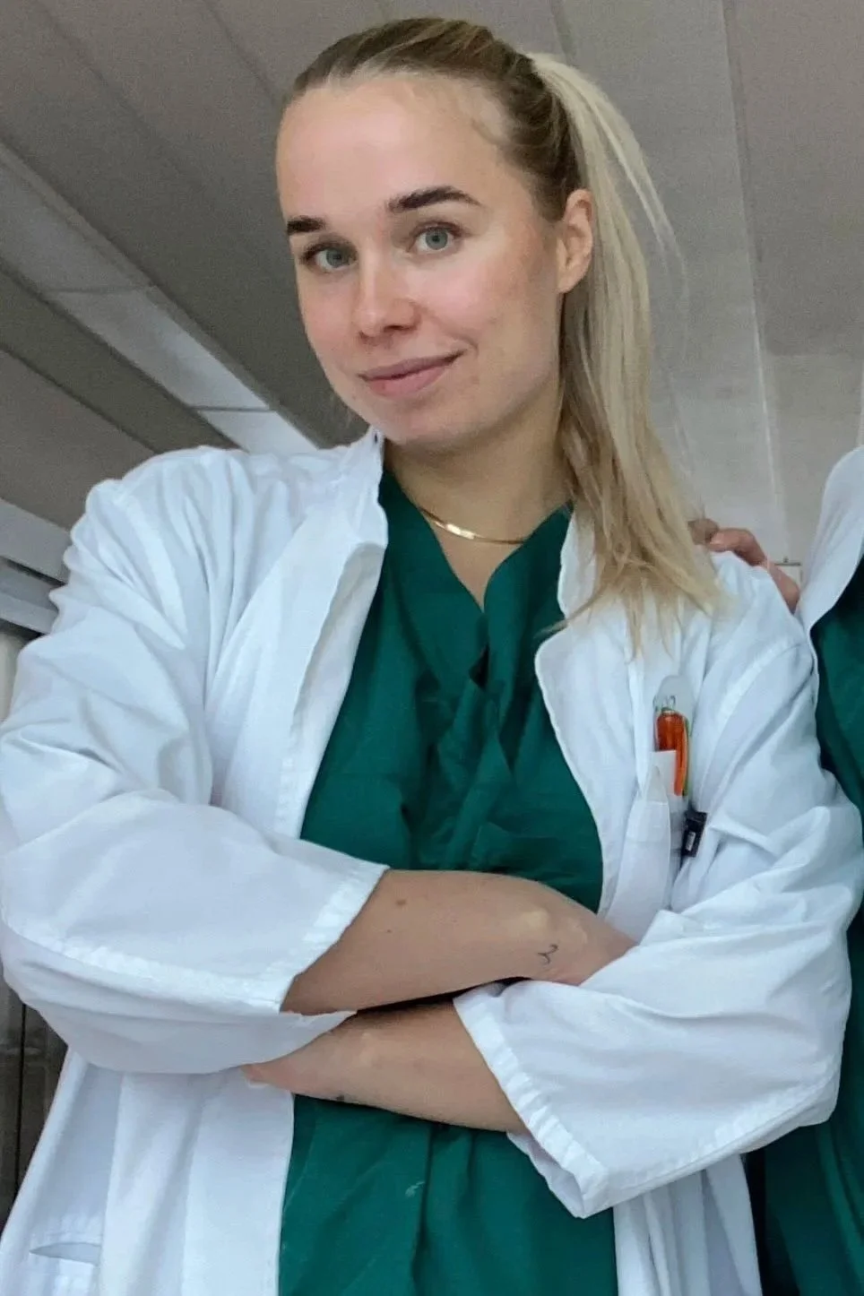 A young woman in medical scrubs and a white lab coat, smiling with arms crossed, inside a clinical or hospital setting.
