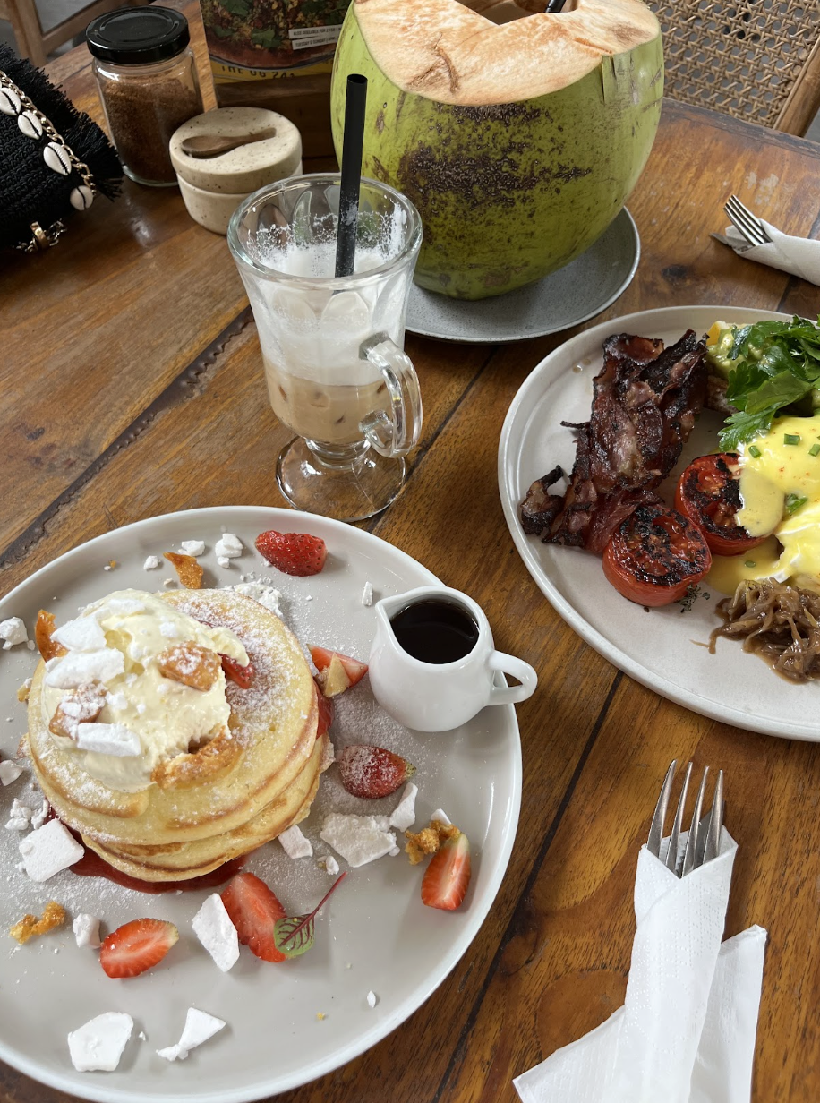 A wooden table with a plate of pancakes topped with strawberries, whipped cream, and powdered sugar, served with maple syrup. A glass of iced coffee with a straw, a plate of breakfast items including bacon, tomatoes, scrambled eggs, and greens, and a large green coconut with a straw are also on the table.