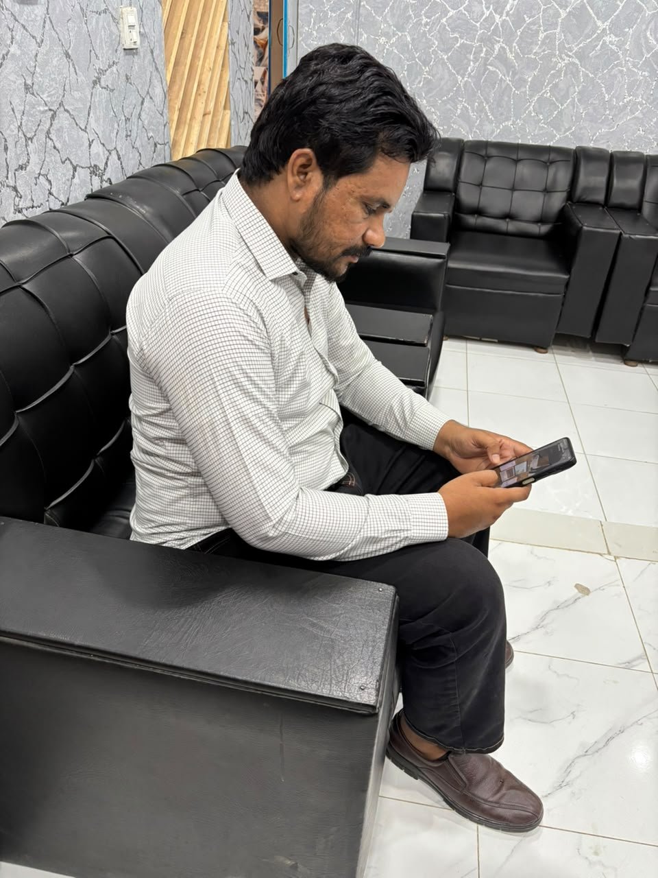 A man with dark hair and beard sitting on a black leather couch, looking at his smartphone in a waiting area with gray textured walls and marble flooring.