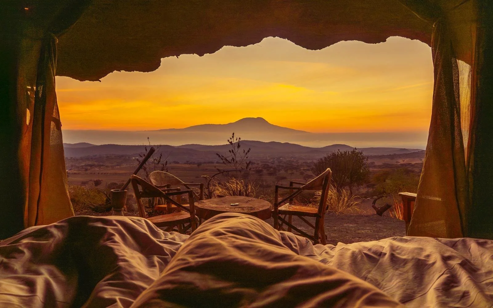 A sunset view from inside a tent showing trees and a mountain in the distance, with a bed and outdoor chairs in the foreground.