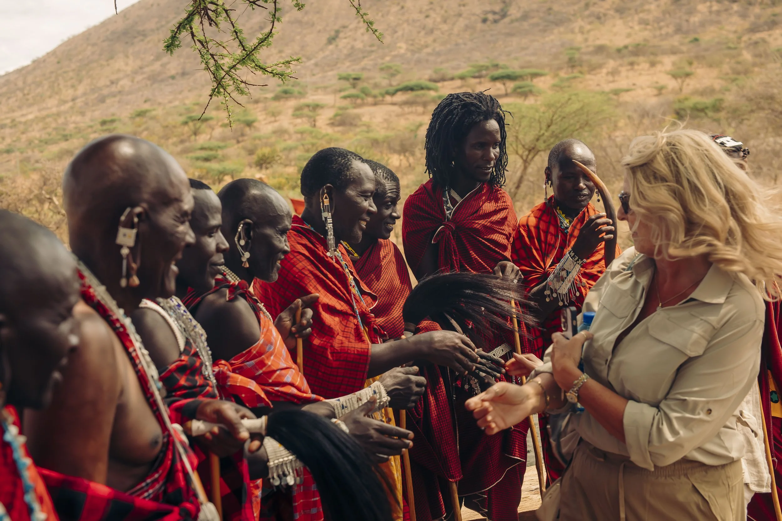A group of indigenous African people, mostly men, dressed in traditional red and black patterned clothing and jewelry, are seated outdoors in a semi-circle, interacting with a blonde woman in a beige shirt, in a dry, hilly landscape with sparse trees.