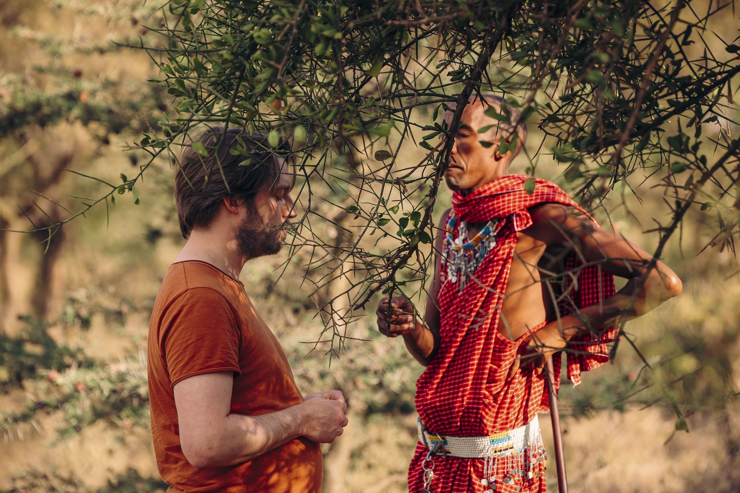 Two men are standing outdoors behind a bush. One man, with long brown hair and a beard, is wearing an orange shirt. The other man, with short hair and facial features suggesting African heritage, is dressed in traditional attire with a red checkered cloth and beaded accessories.