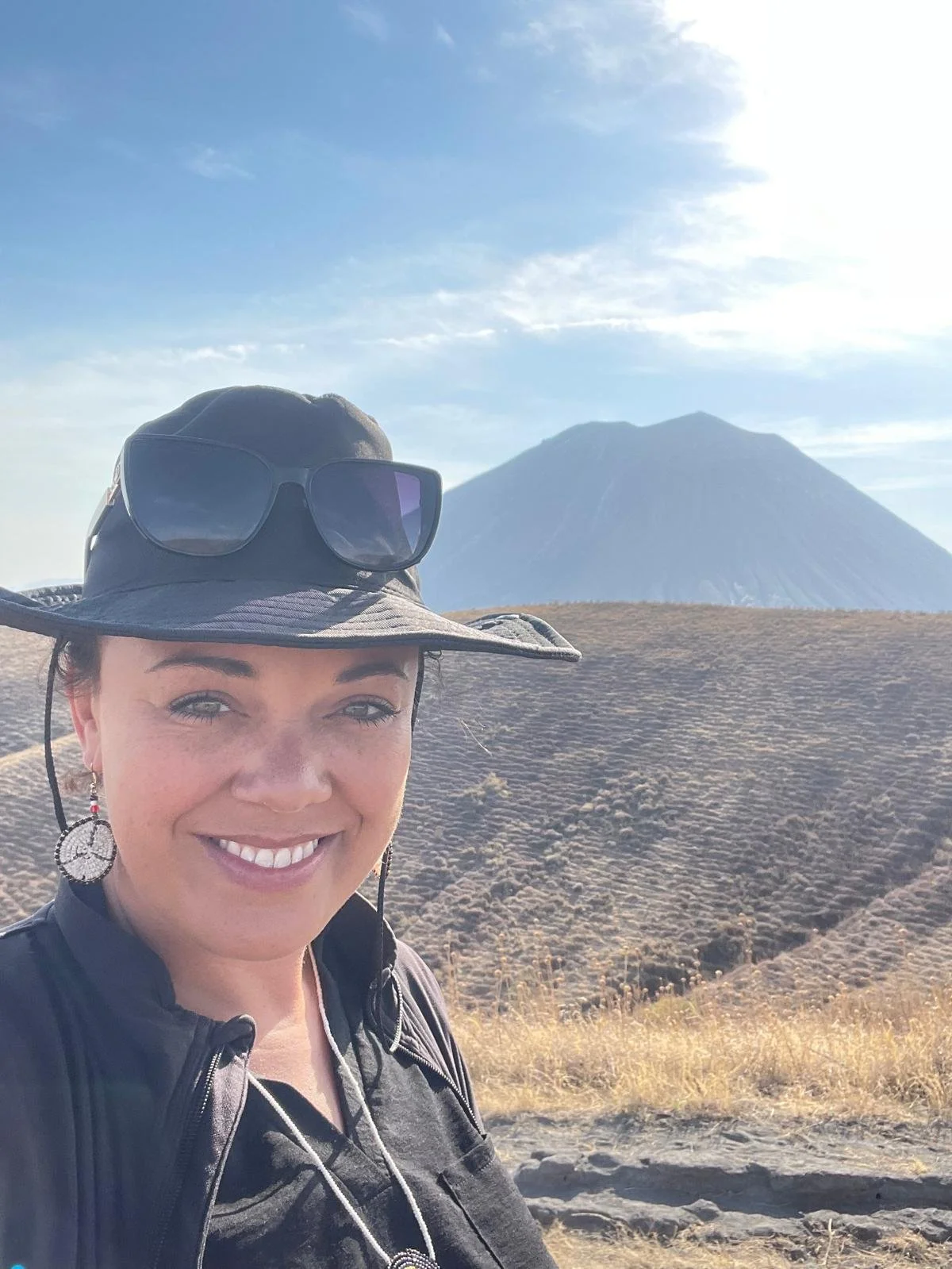 A woman with a wide-brimmed hat and sunglasses on her head, smiling in front of a mountain with a clear sky.
