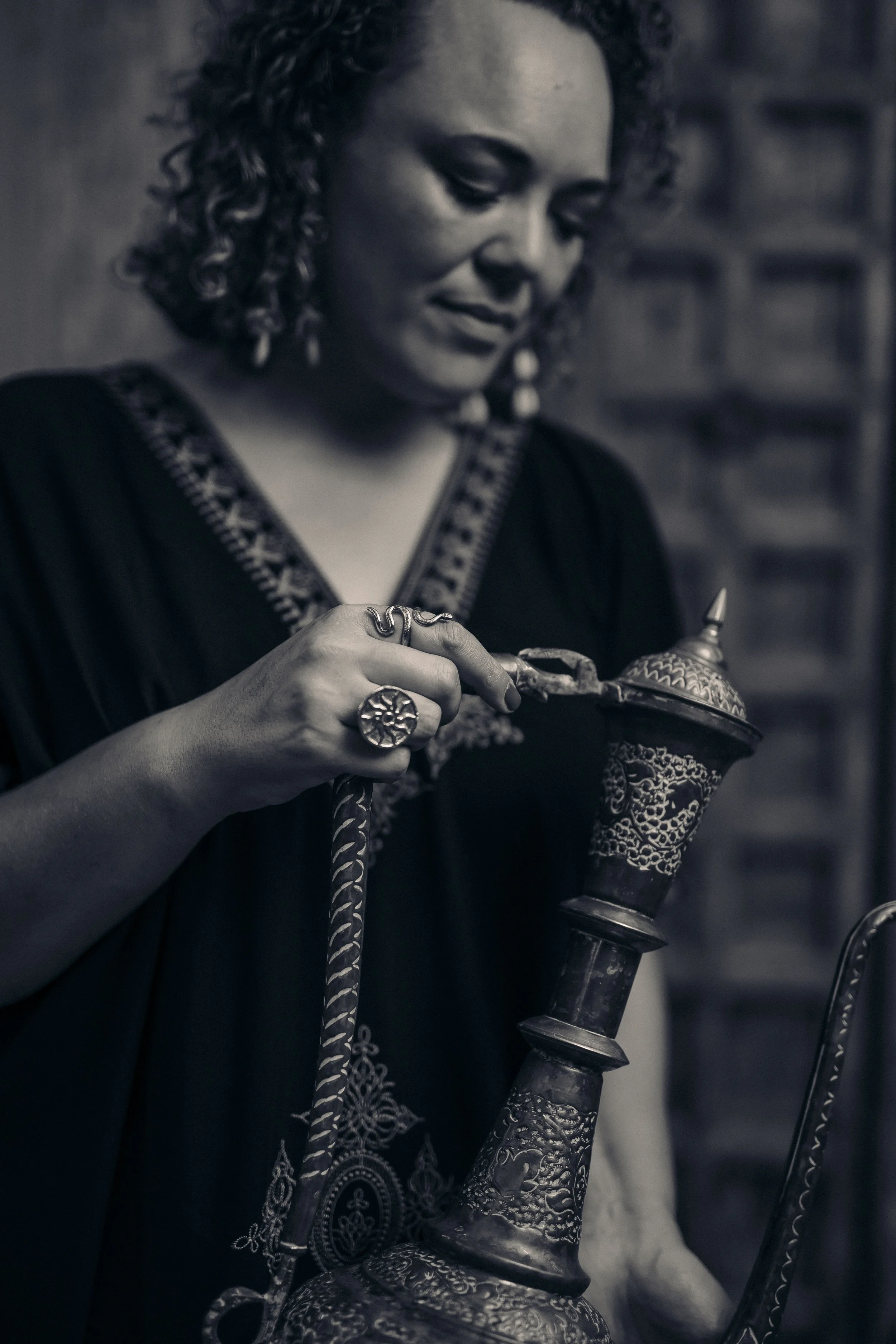 A woman with curly hair, wearing earrings and rings, is holding a traditional decorative metal object with intricate patterns and a pointed tip. The image is in black and white. The Temple Codes