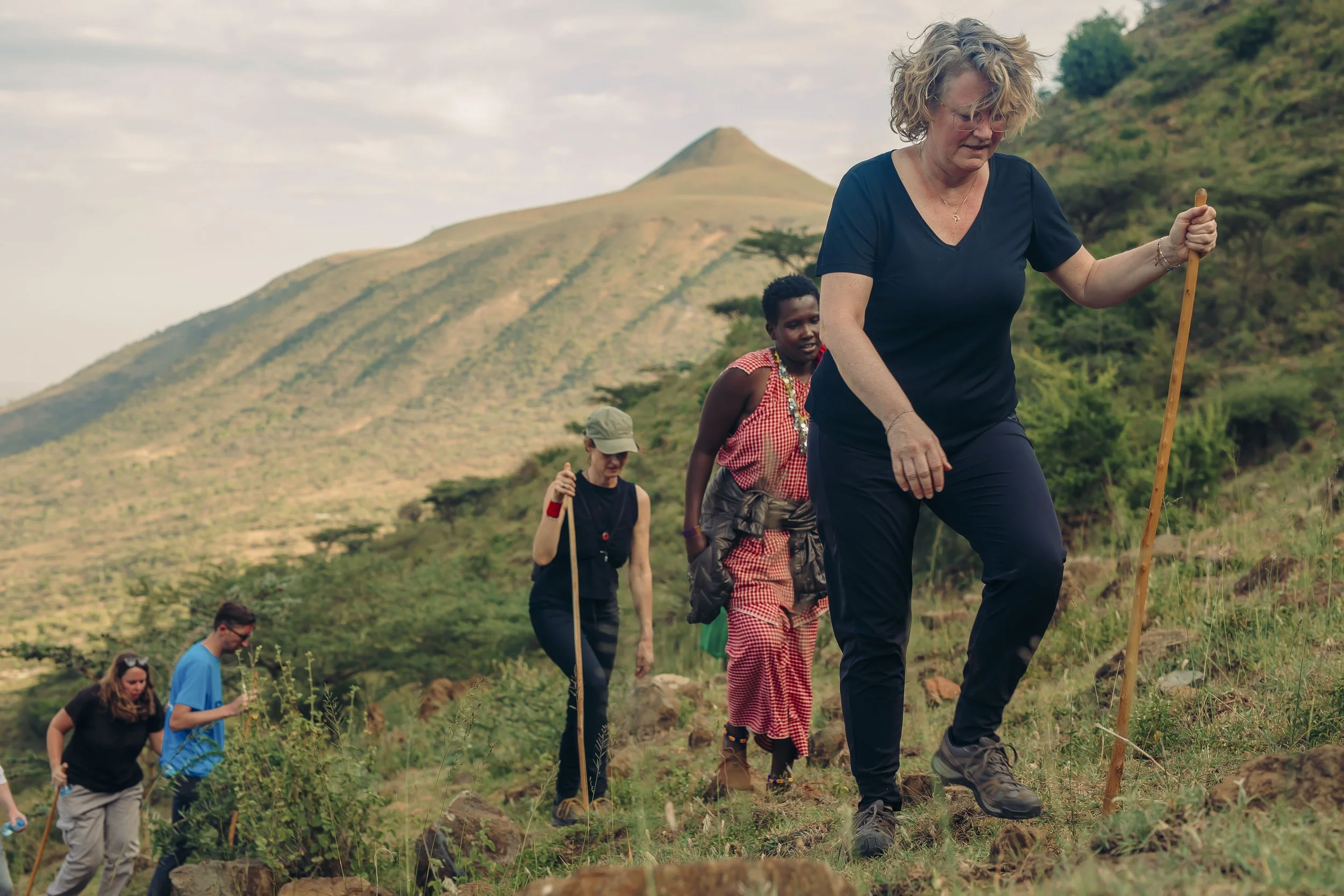 A group of people hiking on a hillside with a mountain in the background. The group includes a woman and children, walking in single file on a trail surrounded by green vegetation.
