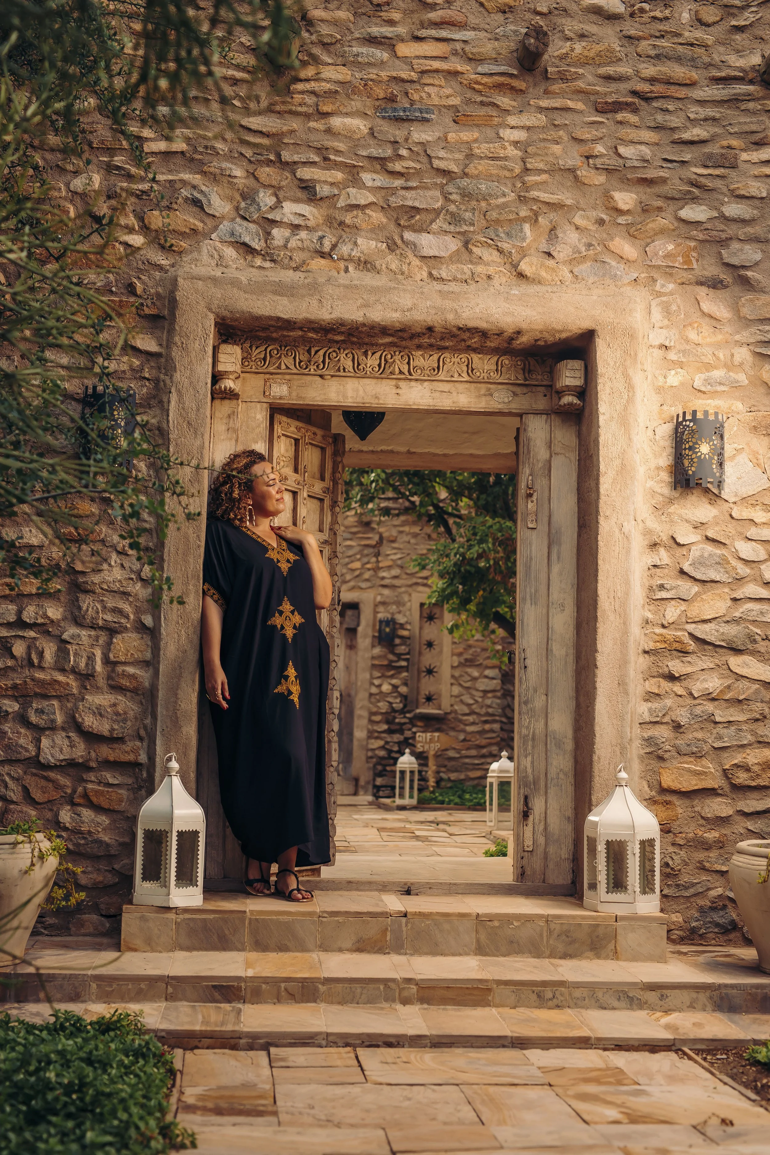 A woman standing at a stone doorway in a rustic setting, wearing a black dress with gold embroidery, looking to the side with her hand on her shoulder. There are white lanterns on the ground and greenery around.