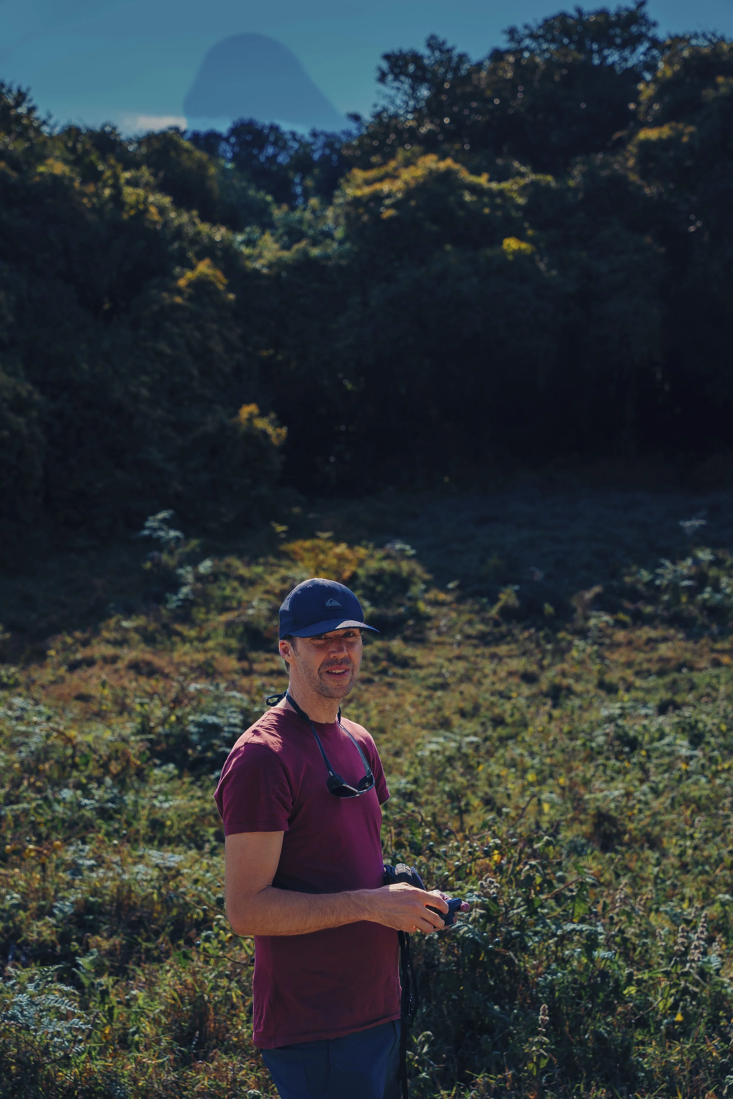 A mountain-guide in a maroon t-shirt and navy cap standing outdoors in a grassy, shrub-filled area with trees and a blue sky in the background.