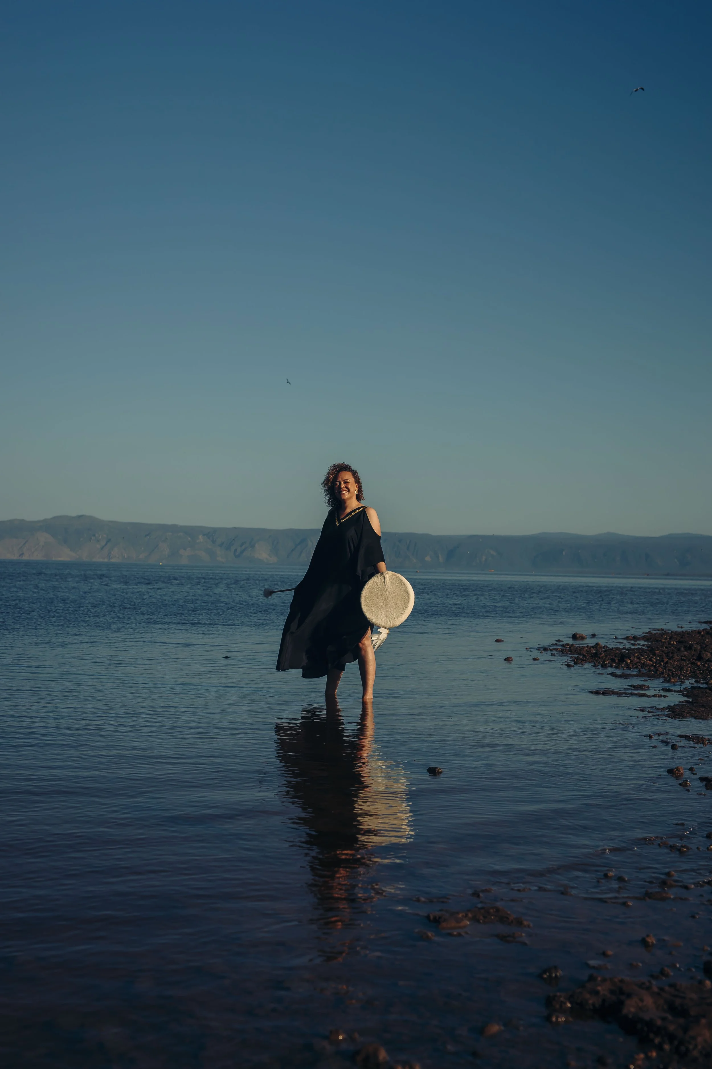 A woman standing in shallow water at the beach, holding a tambourine in one hand, smiling, with mountains in the background and a clear sky above.