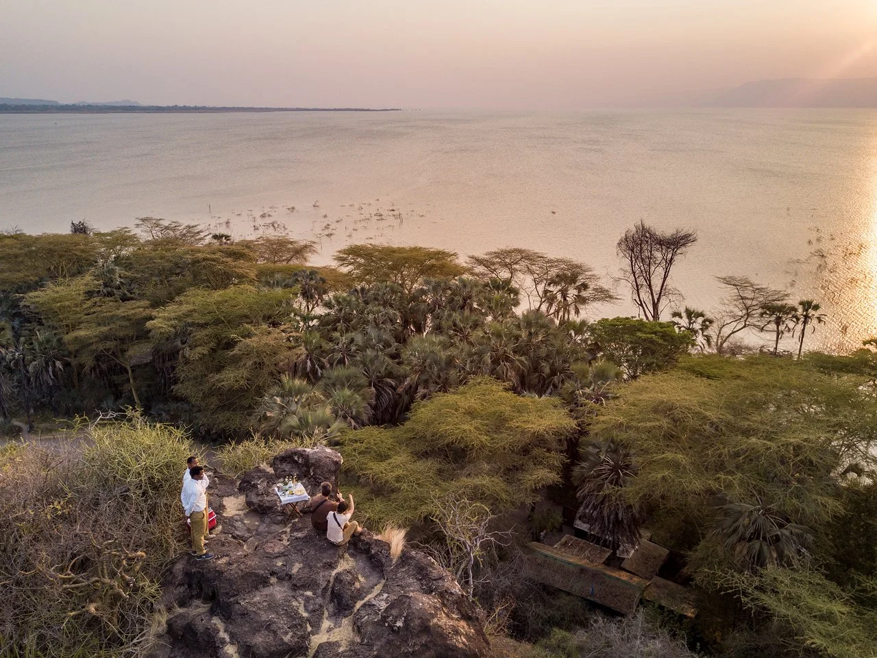 A scenic view of a group of people sitting on rocky terrain overlooking a lush forest with tall trees and a large body of water, Lake Eyasi, in the background at sunset.