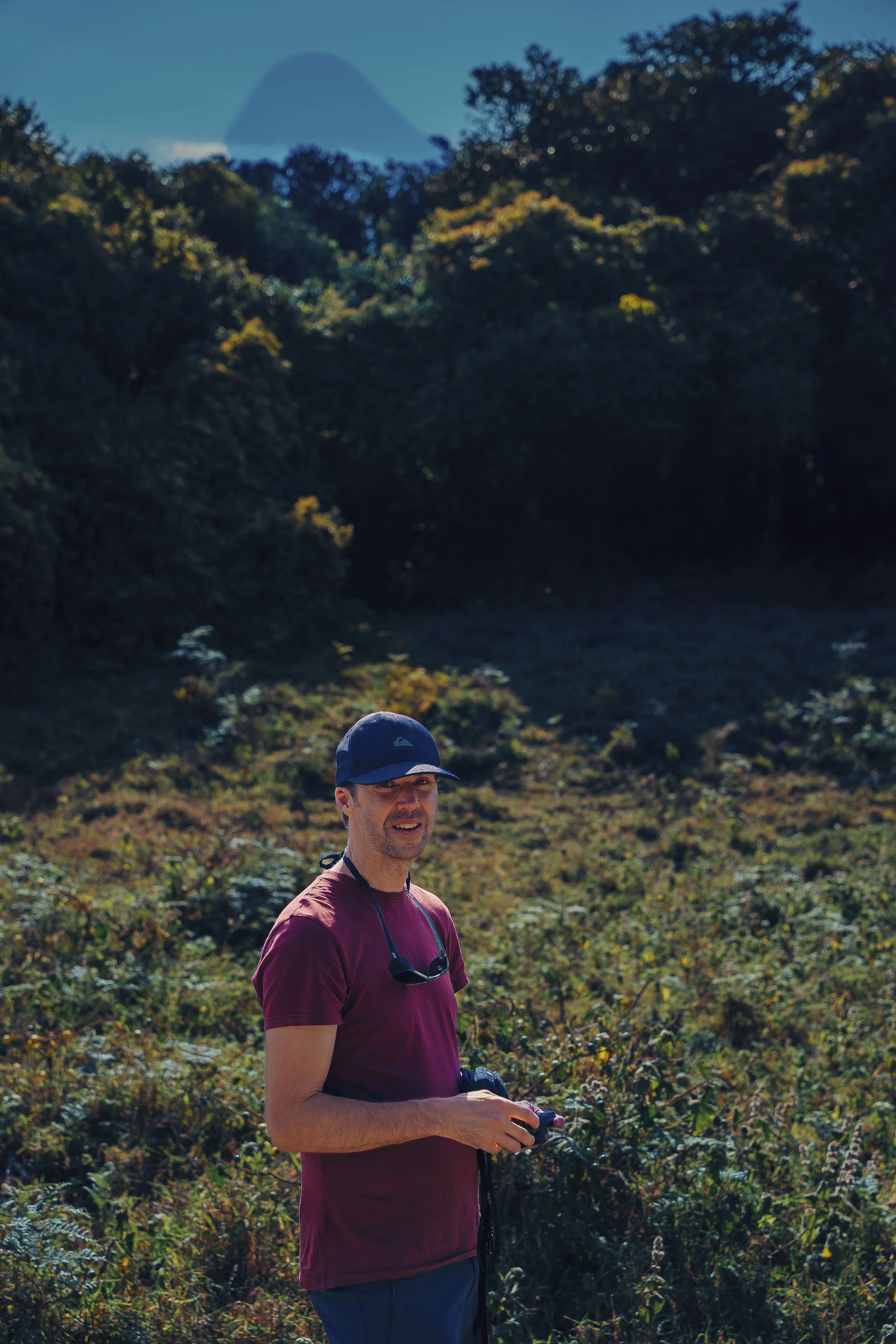 A man with a cap and sunglasses hanging from his shirt stands outdoors in a natural setting with dense vegetation and trees, holding a camera or device in his hands. In the background, a mountain is visible under a clear blue sky.