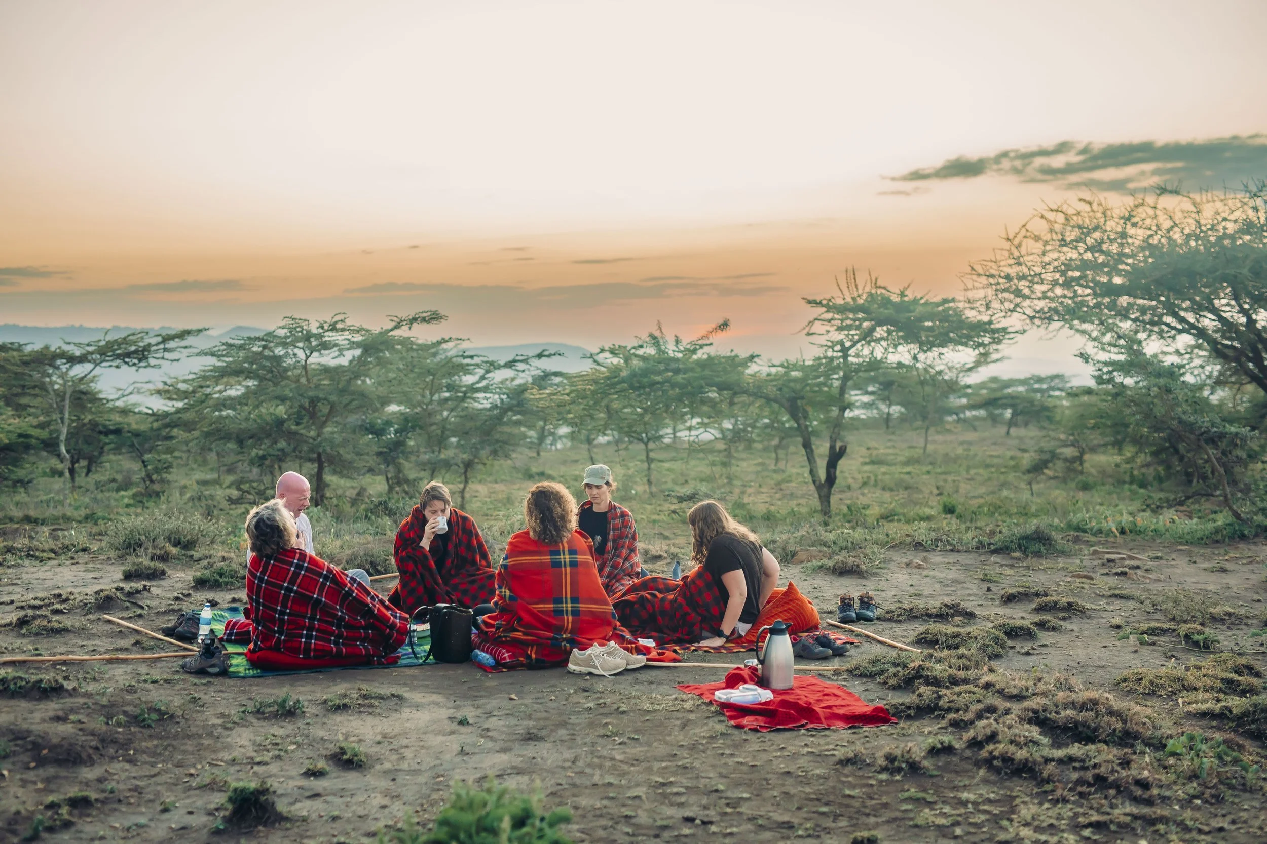 A group of people sitting on blankets in a semi-circle outdoors during sunset, surrounded by sparse trees and a mountain range in the distance, with some drinking and talking.