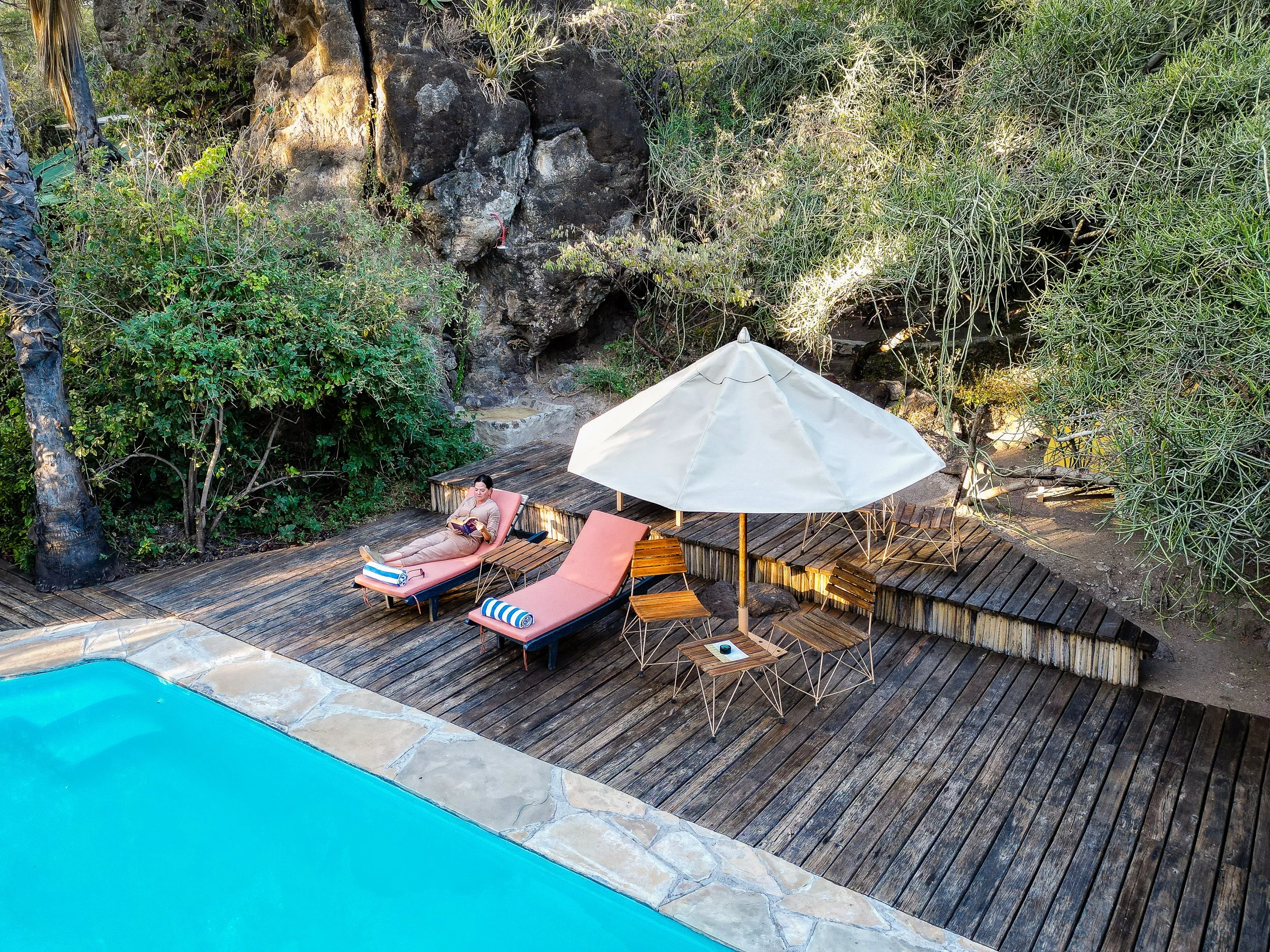 A person relaxing on a pink lounge chair beside a swimming pool, surrounded by a wooden deck, with a large white umbrella and lush greenery including trees and bushes.