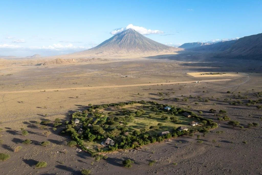 A small green village with several houses and trees is situated in a barren arid landscape, with a large mountain in the background under a clear blue sky.