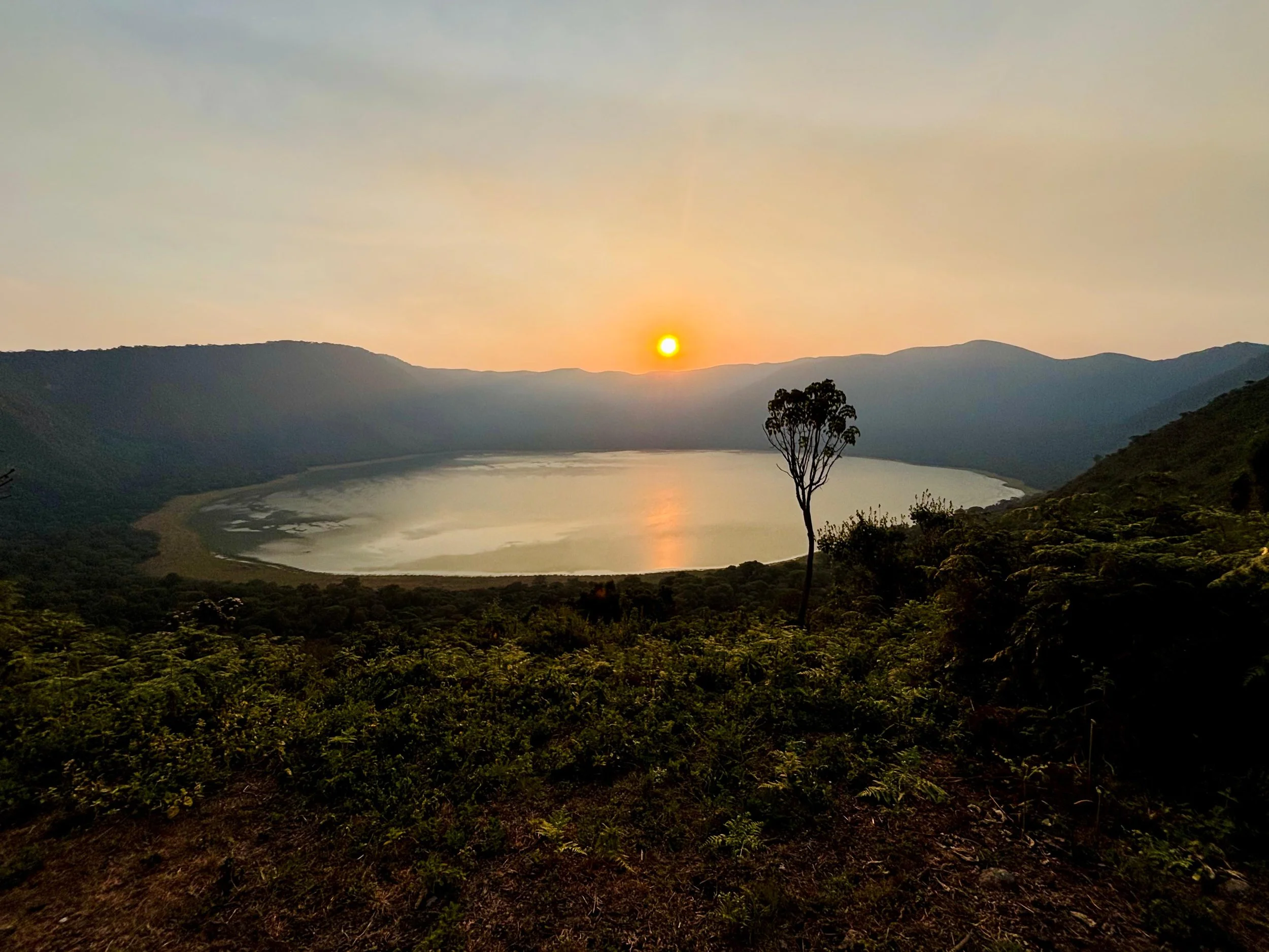 Sun setting over Empakaai Crater's lake surrounded by hills and trees.