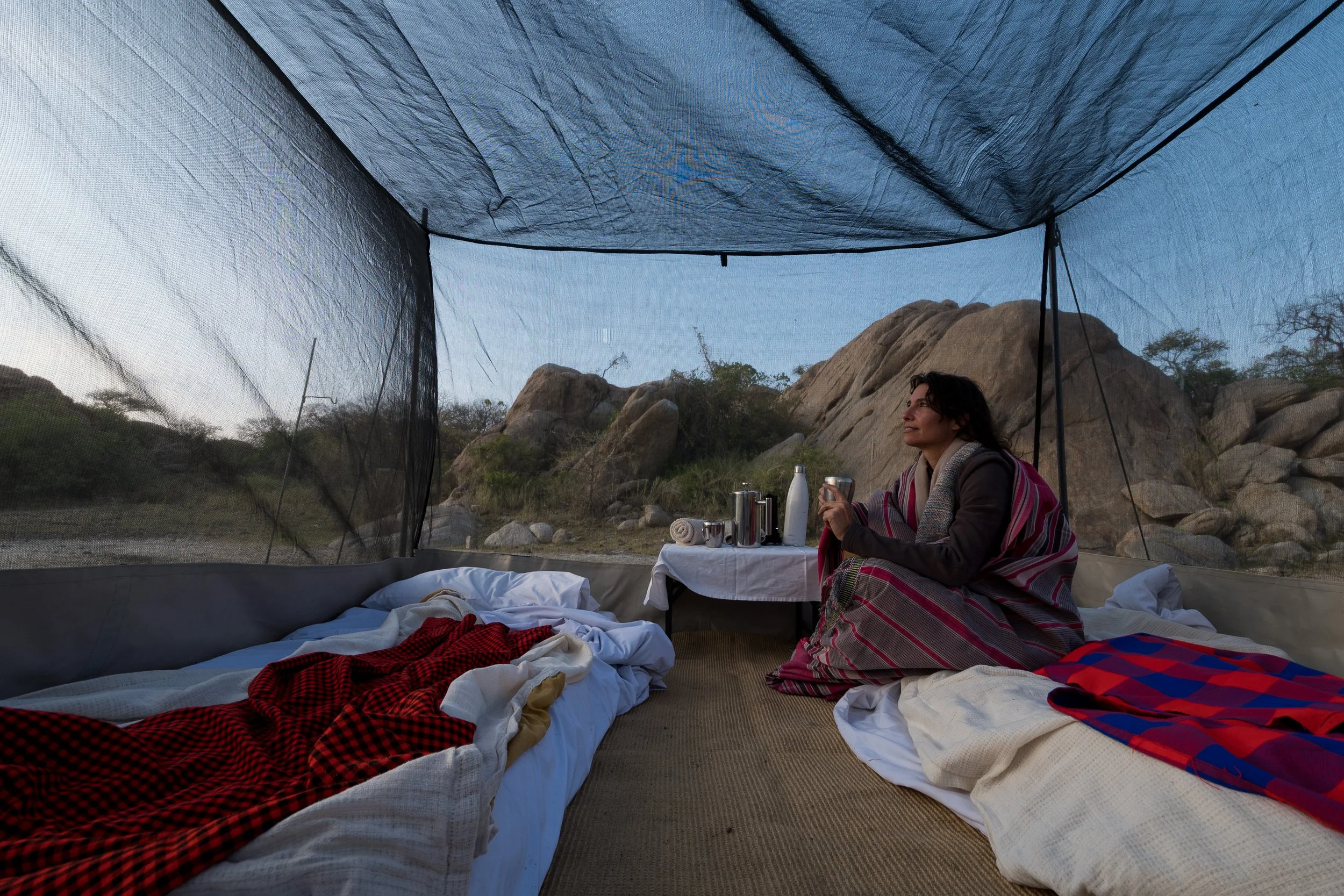 Woman sitting inside a camping tent outdoors, fly camping, with a rocky landscape and trees visible outside, holding a cup and smiling. The tent's interior has a bed with blankets and items on a small table.