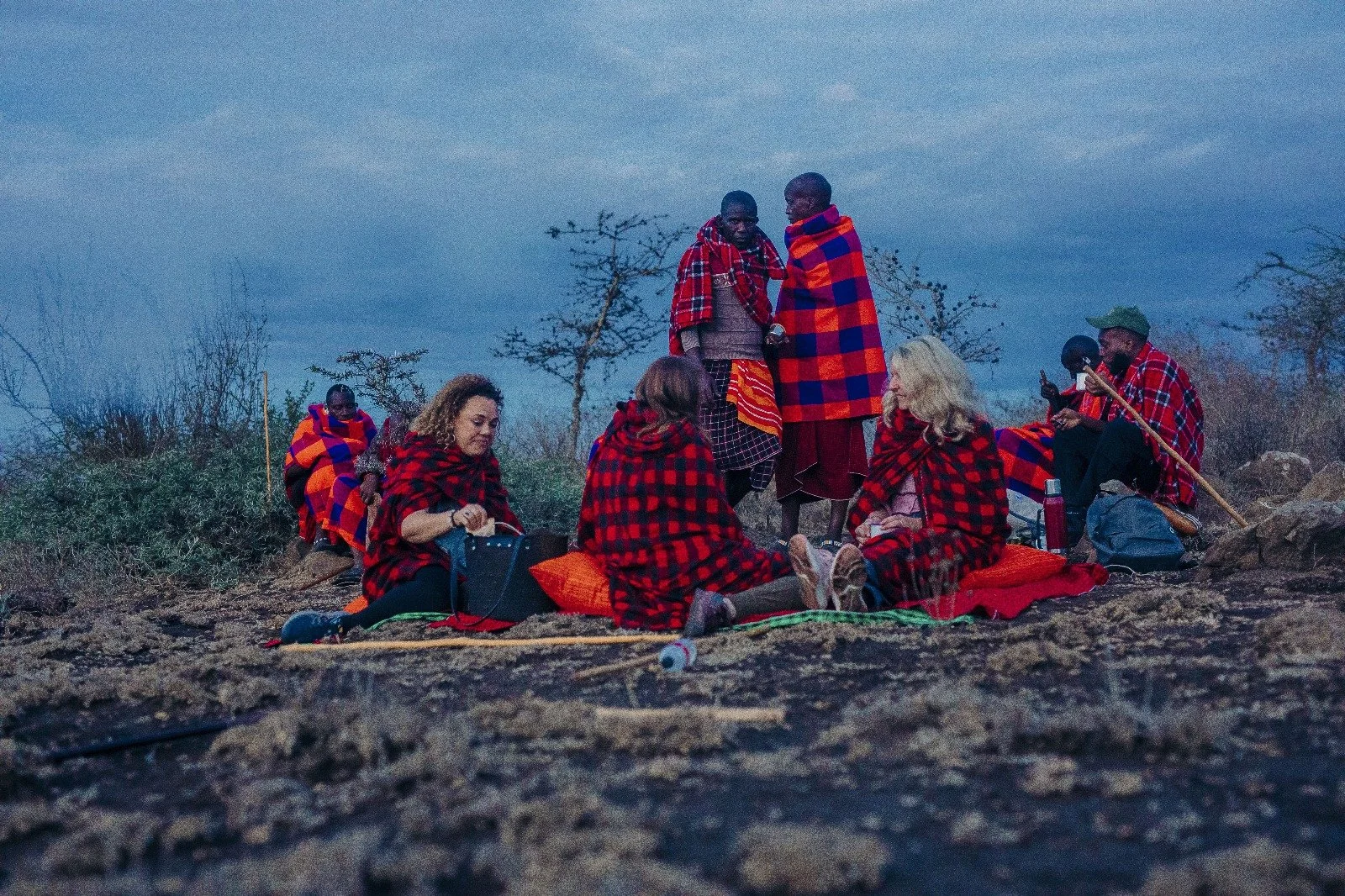 A group of people sitting and standing outdoors on a rocky terrain, wrapped in red and black plaid blankets, during dusk or dawn, with plant life and leafless trees in the background.