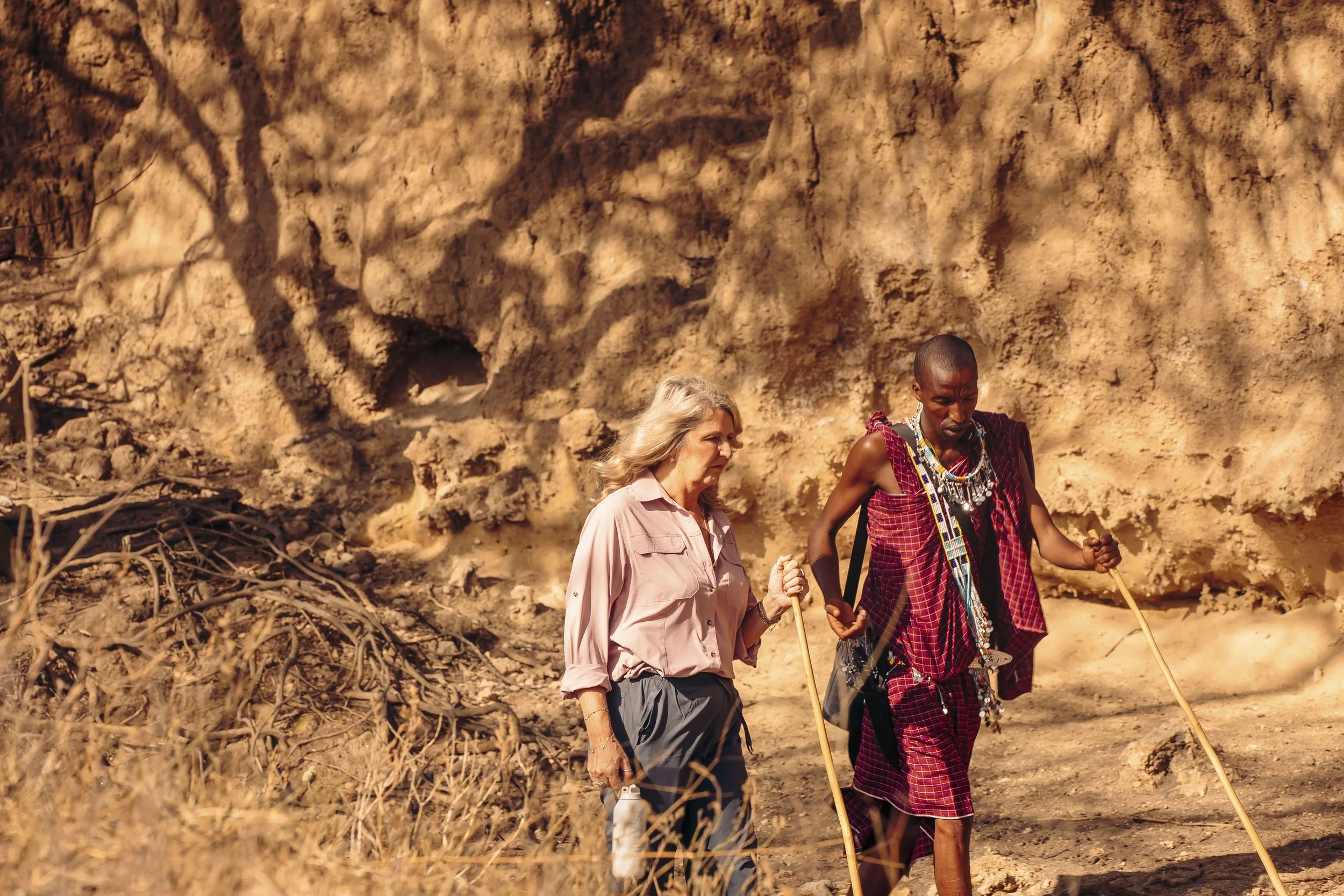 Two women, one with blonde hair and light skin, and the other with dark skin and short hair wearing traditional Maasai attire, walking together with sticks in hand along a dirt path against a rocky hillside.