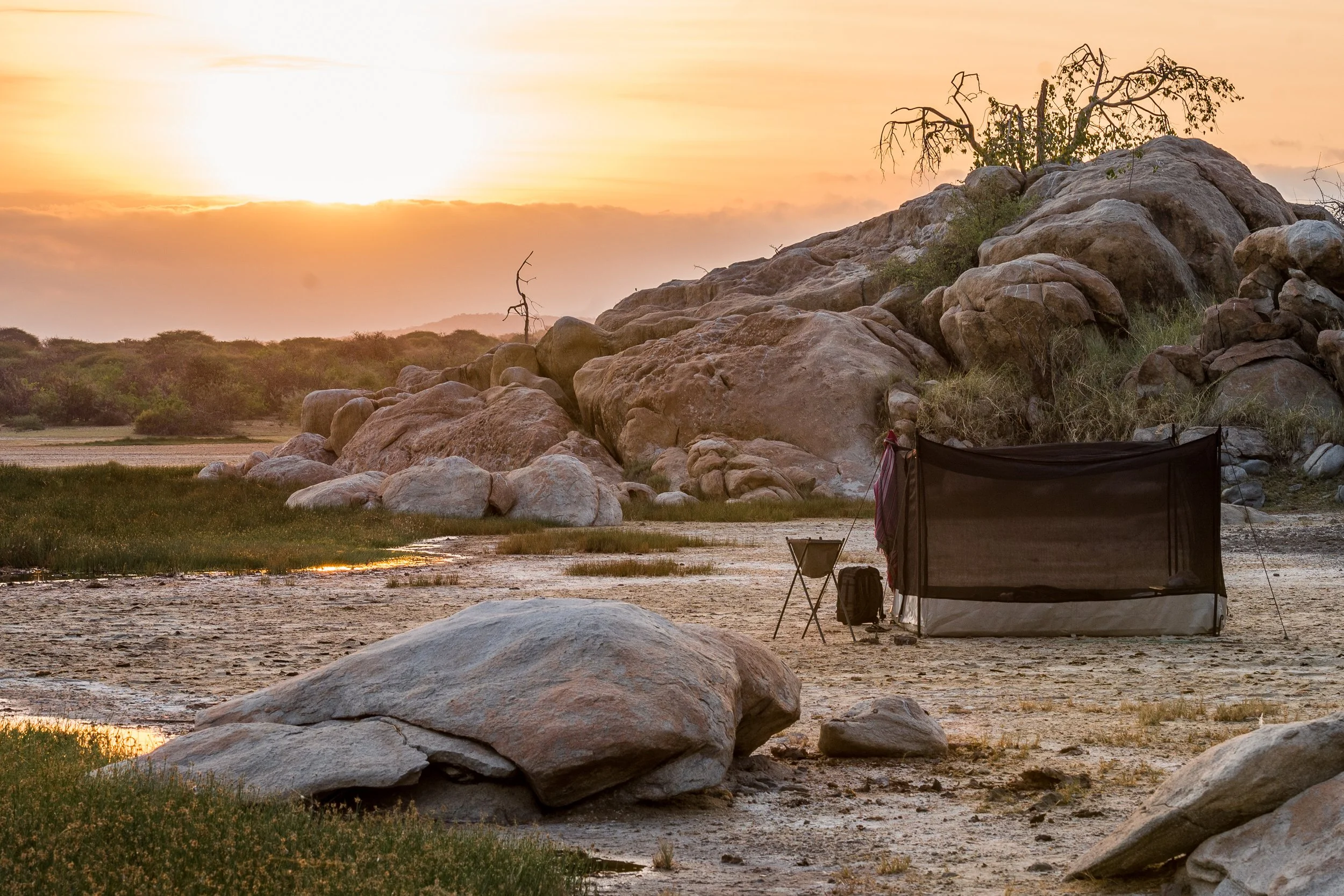 Sunset over a rocky landscape with sparse trees, a camping tent, and outdoor chairs on a sandy ground.