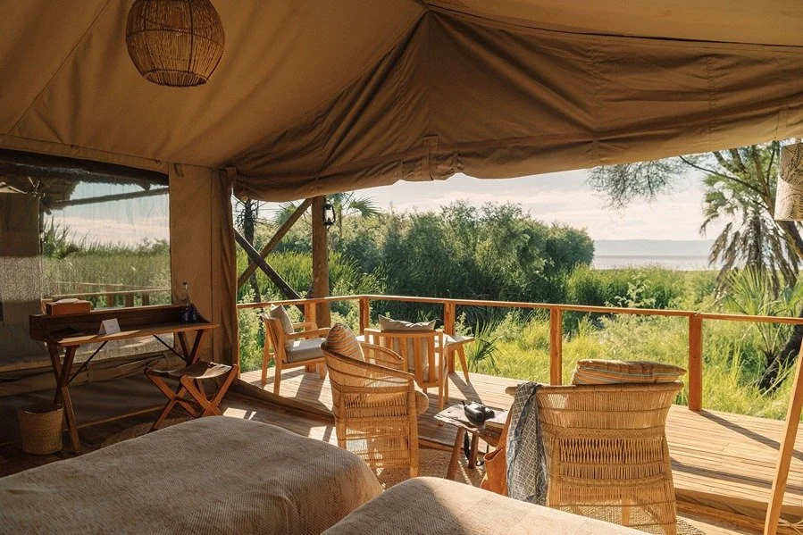 Interior of a tent with beds, wicker chairs, and a wooden deck overlooking lush green vegetation and water in the background.