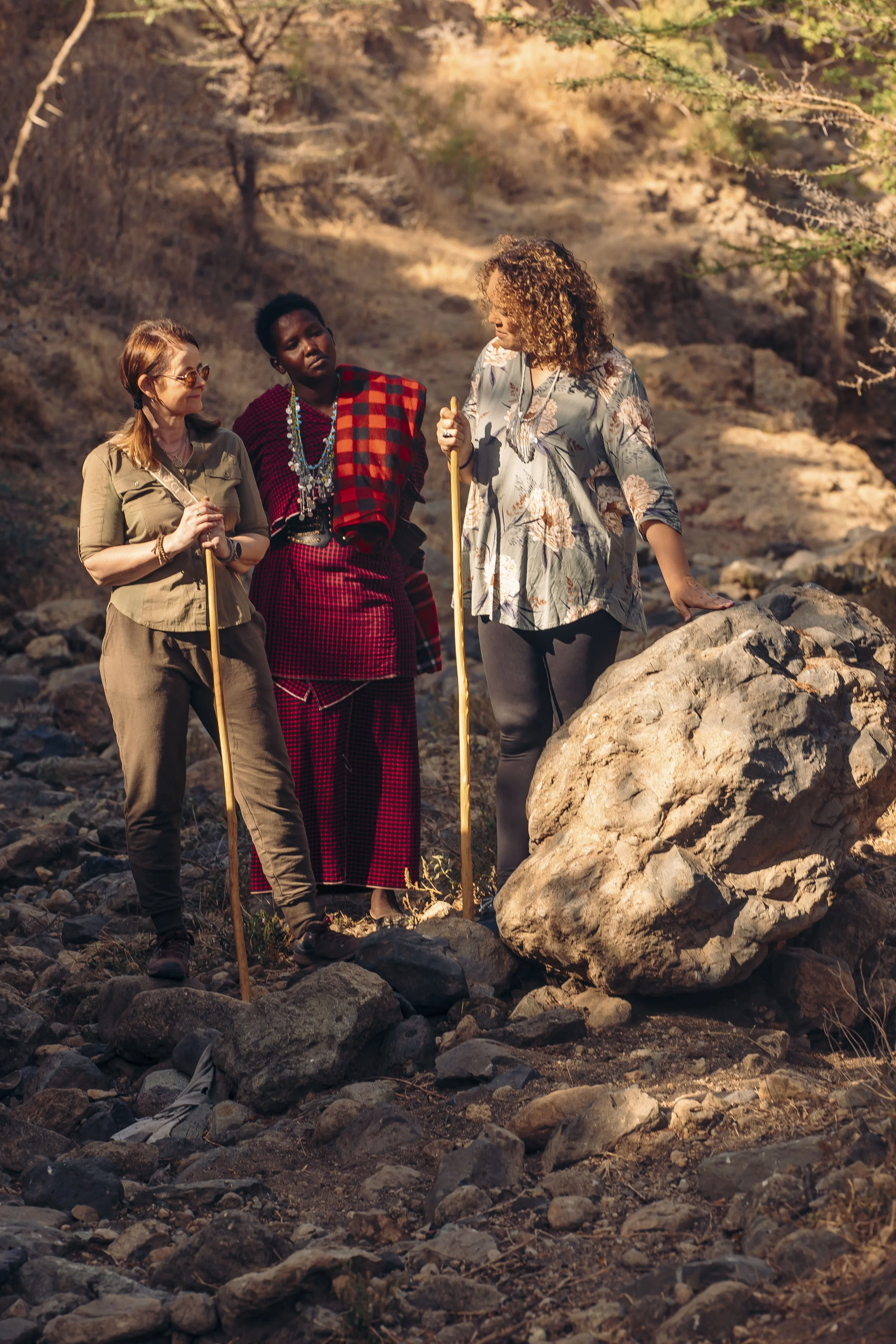 Three women standing outdoors on rocky terrain, engaging in conversation during late afternoon or early evening, with dry bushes and rocks in the background.