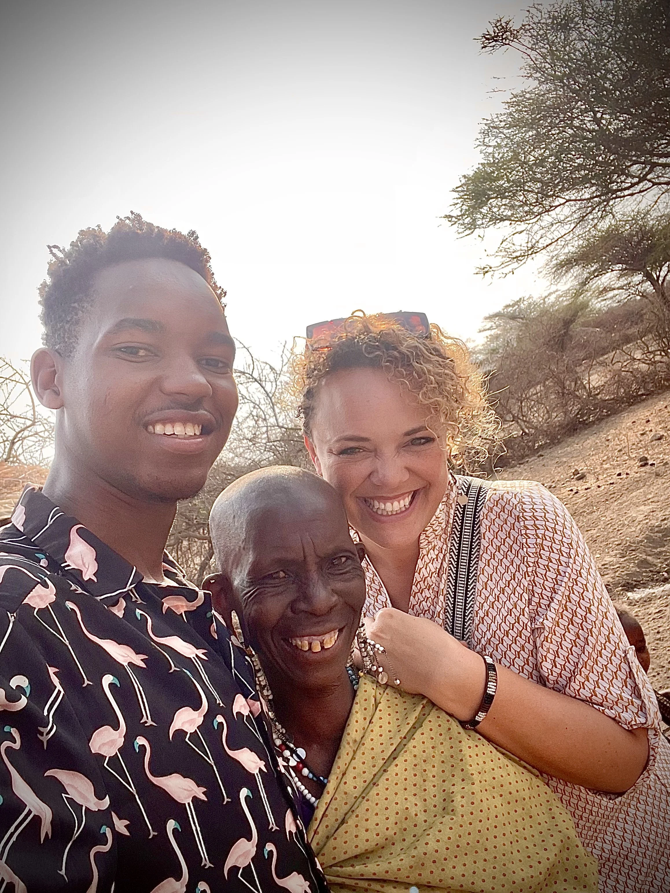 Four people smiling and embracing each other outdoors in a desert-like environment with dry trees, during sunset or sunrise.