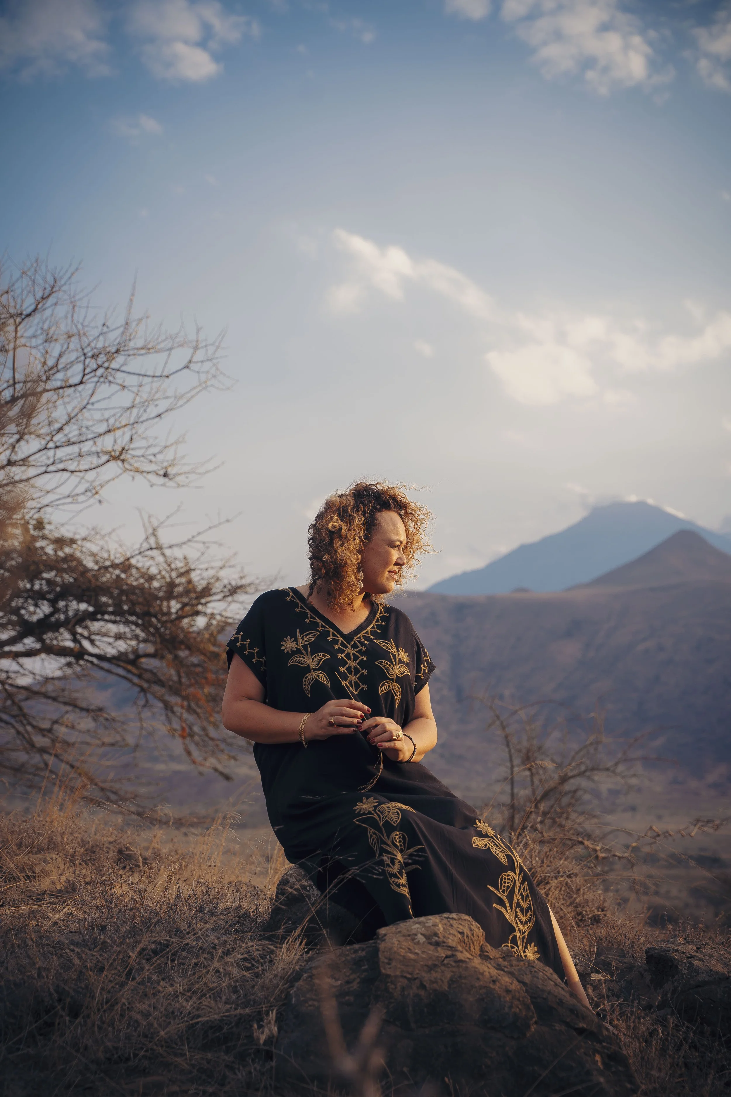 A woman with curly hair wearing a black dress with floral embroidery, sitting on a rock in an arid landscape with mountains and a blue sky with scattered clouds in the background.