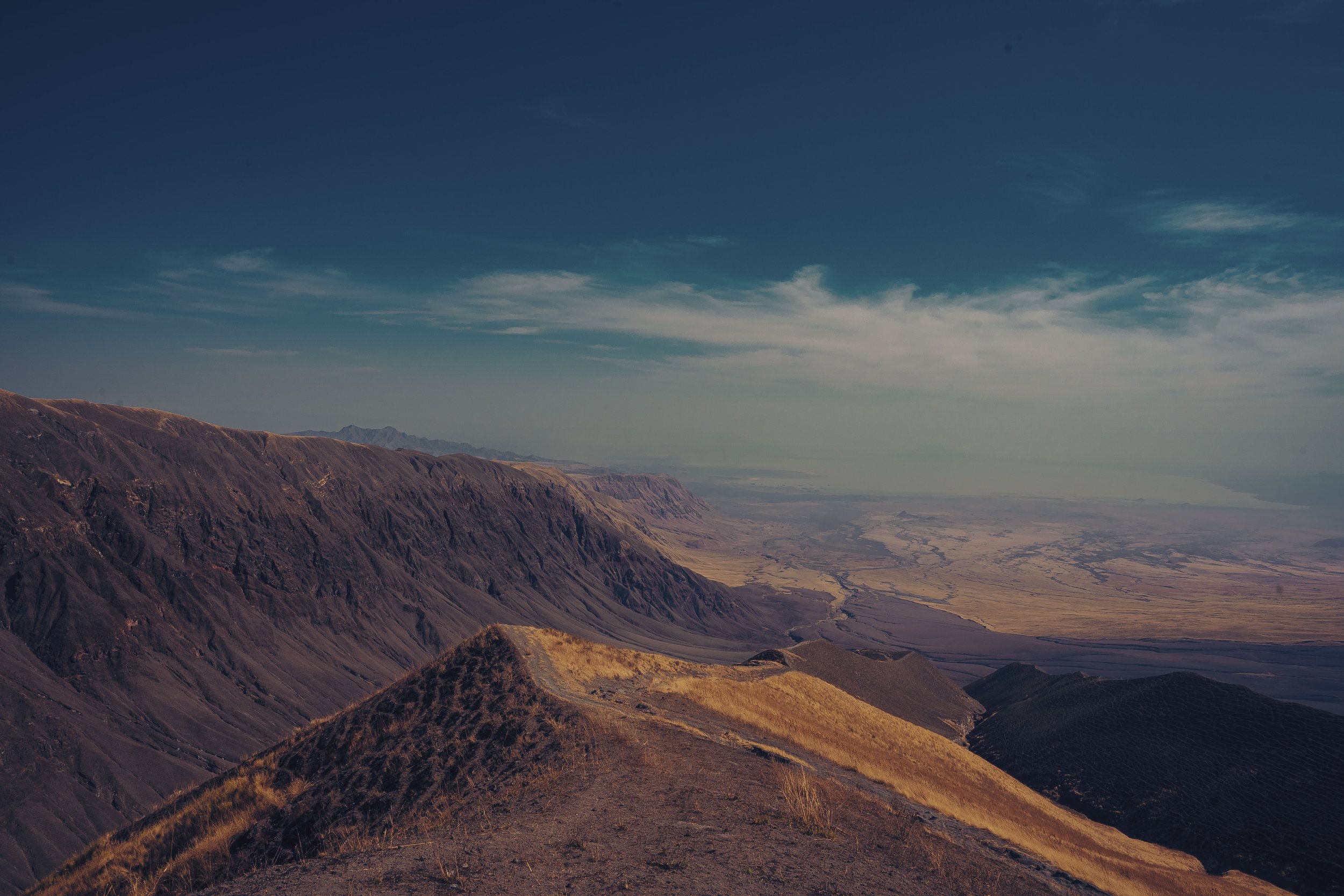 Wide view of arid, rugged mountains with a broad valley and dry riverbeds beneath a partially cloudy sky.