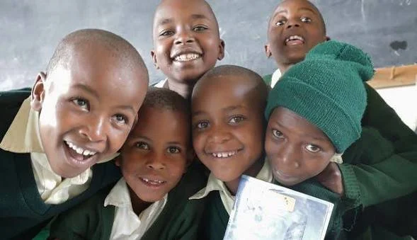 Six smiling African children, some wearing green and cream uniforms, gathered closely together in a classroom.