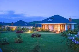 A house with a blue roof illuminated at night, surrounded by a well-maintained garden with bushes and flowers, with mountains and a cloudy sky in the background.