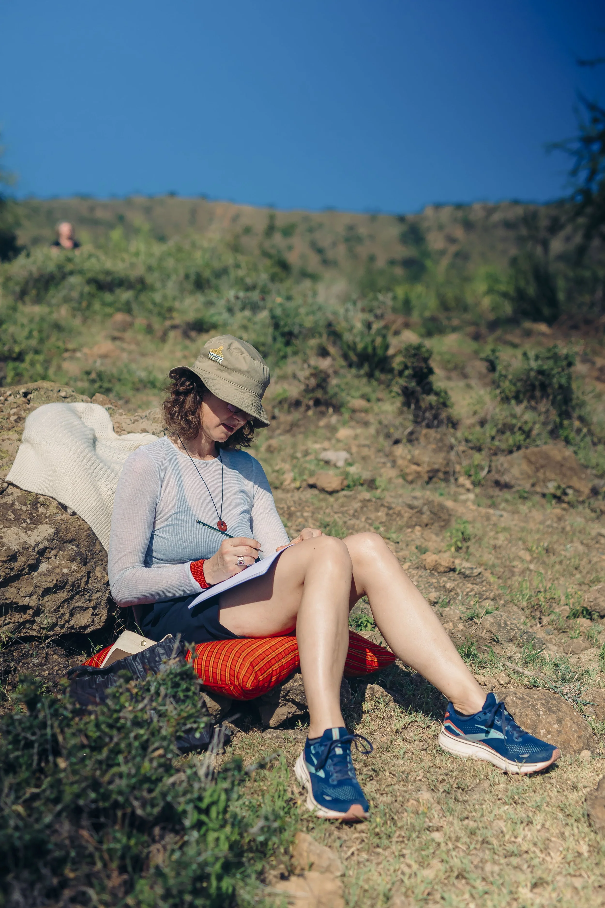 A woman sitting outdoors on a rock, writing in a notebook, wearing a bucket hat, sunglasses, and athletic shoes, with a backpack nearby, in a hilly, green landscape under a clear blue sky.