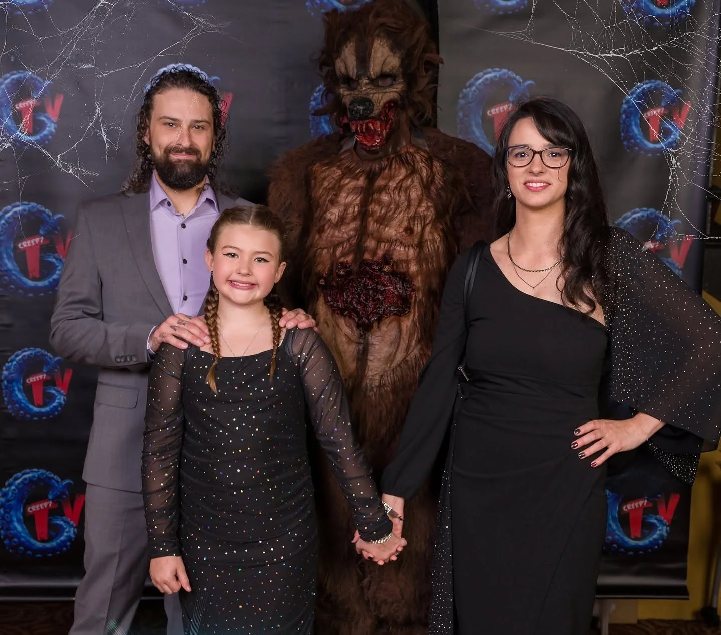 A family with a werewolf costume, standing in front of a backdrop with a creepy TV logo. The family includes a man with long curly hair and beard, a young girl with braids, and a woman with dark hair and glasses, all dressed in festive black outfits. They are smiling and holding hands.