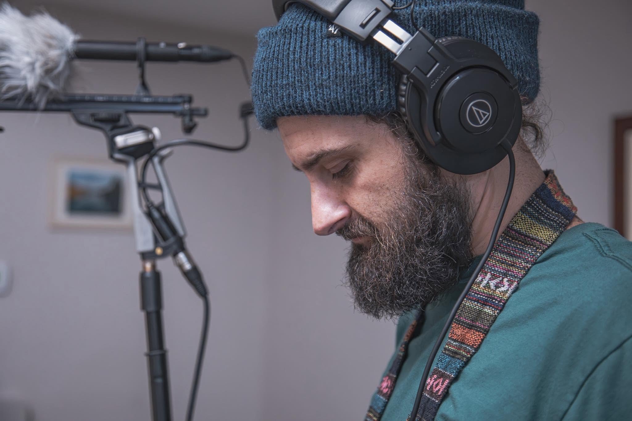Tyler Darkow with a beard and curly hair wearing a blue knit cap and large black headphones, working with audio equipment, with a microphone on a stand in the foreground.