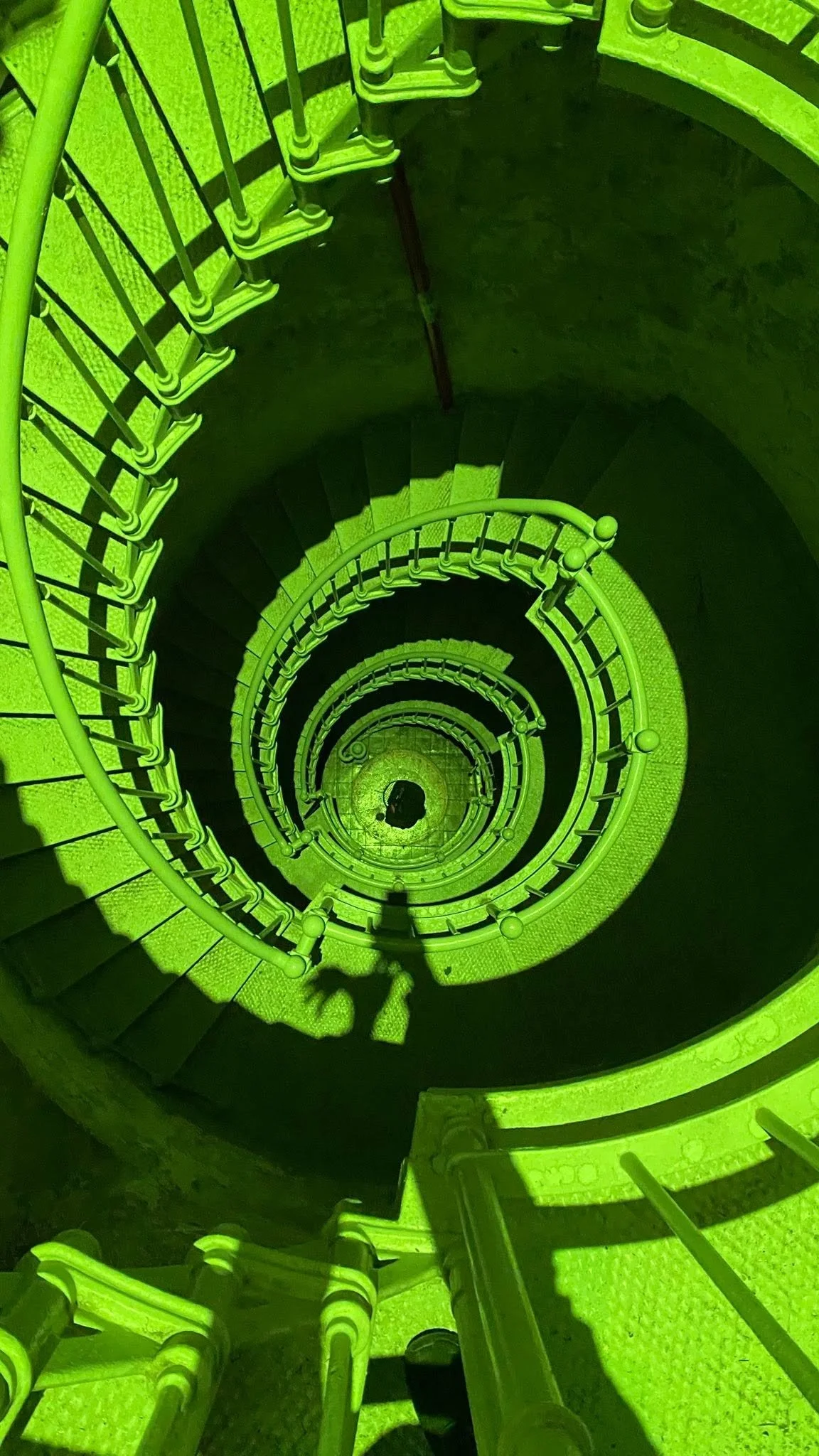 A spiral staircase viewed from the top looking down, with green metal railings and steps, creating a circular pattern.