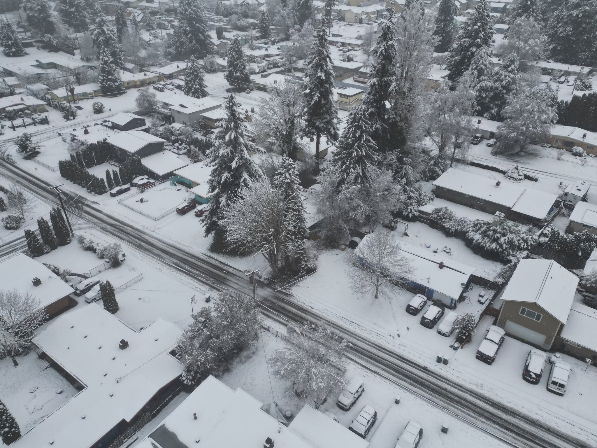 A drone aerial view of a residential neighborhood covered in snow with houses, trees, and parked cars.