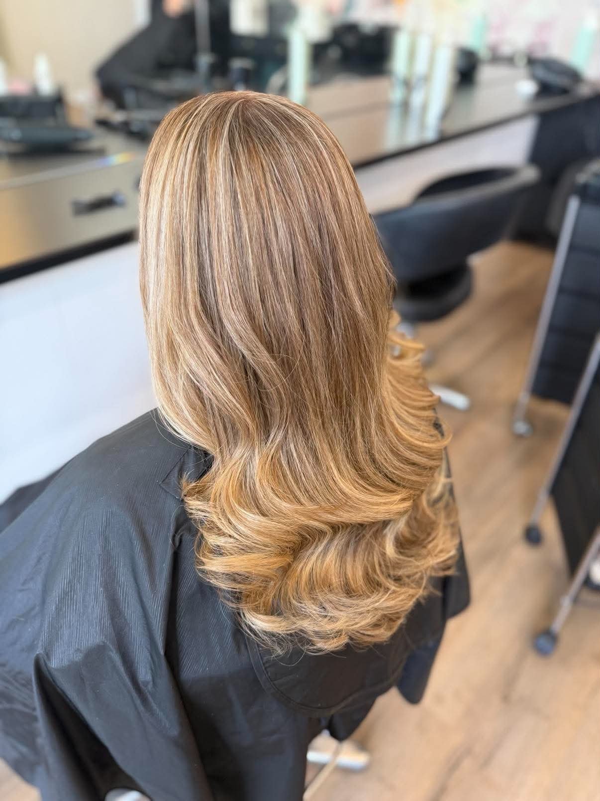 A woman with styled, shoulder-length, blonde hair with soft waves, seated in a hair salon during a haircut or styling session.