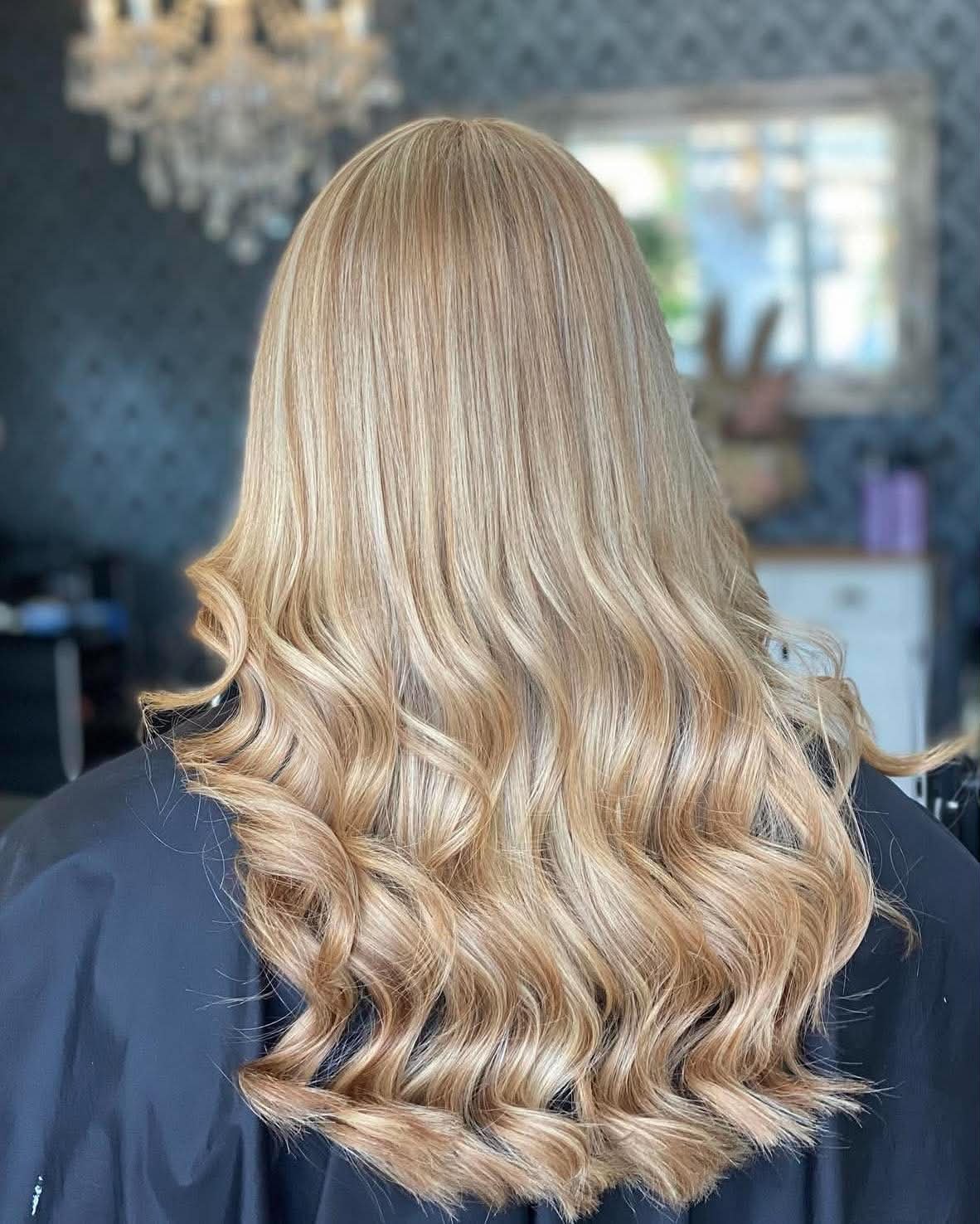 The image shows a woman with long, blonde hair that is styled in loose curls, sitting in a salon or hair studio.